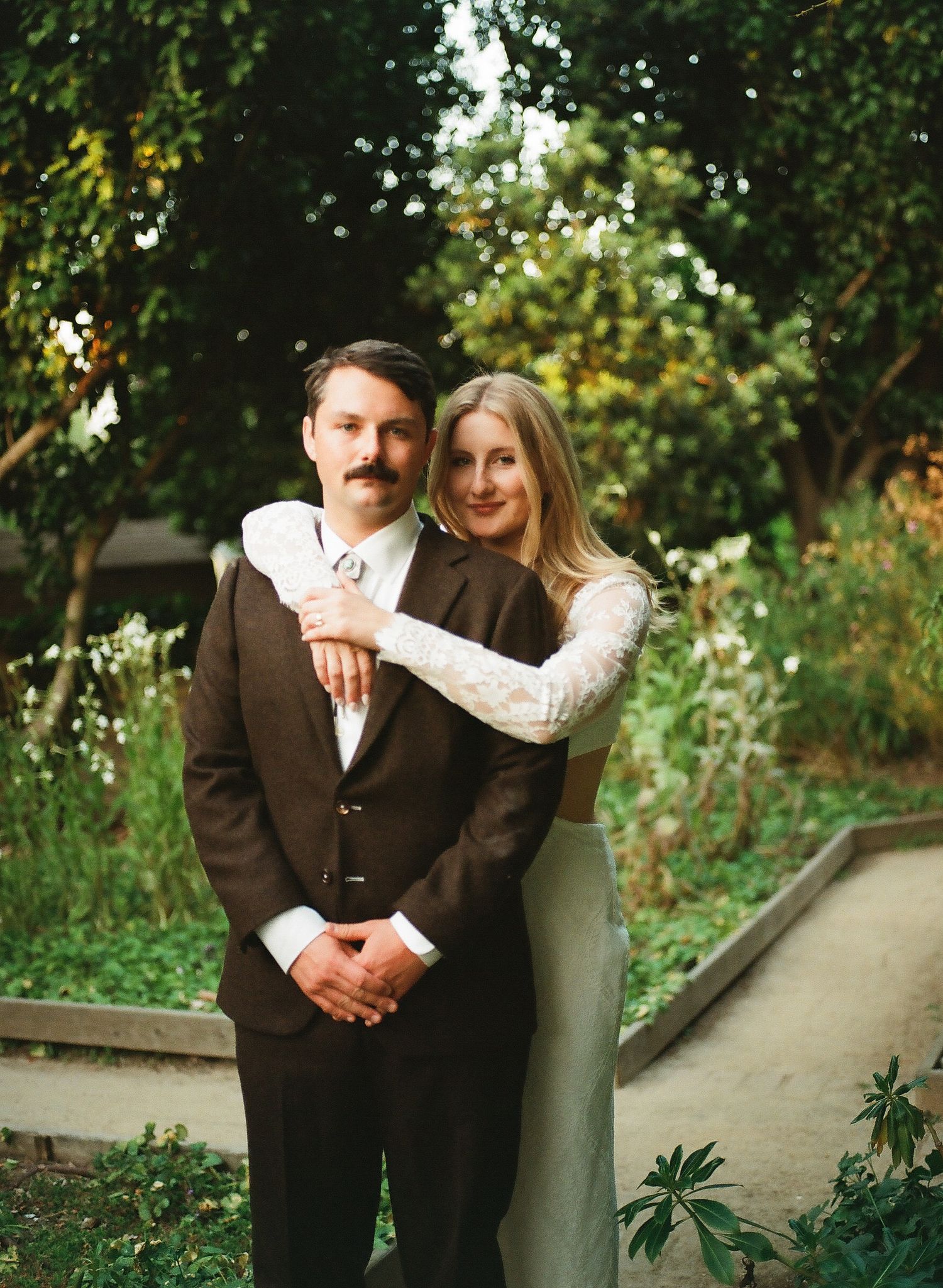 A couple poses in a lush garden, the man in a brown suit and the woman in a white lace top embracing him warmly.