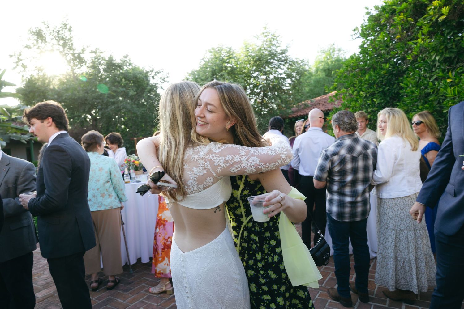 Two women share a warm hug at an outdoor wedding reception surrounded by elegantly dressed guests.
