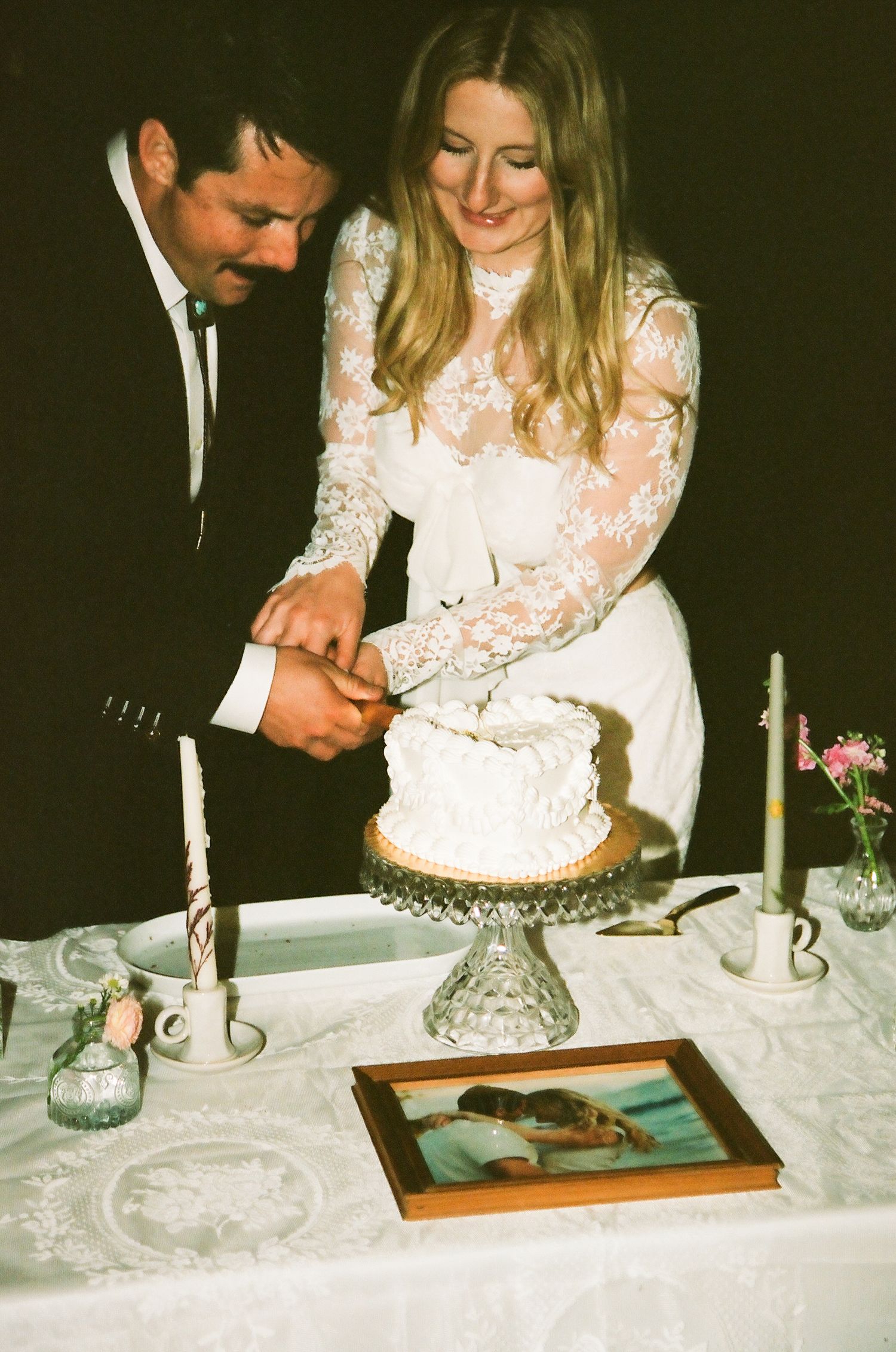 Bride and groom cutting white wedding cake together at reception table with candles and framed photo.