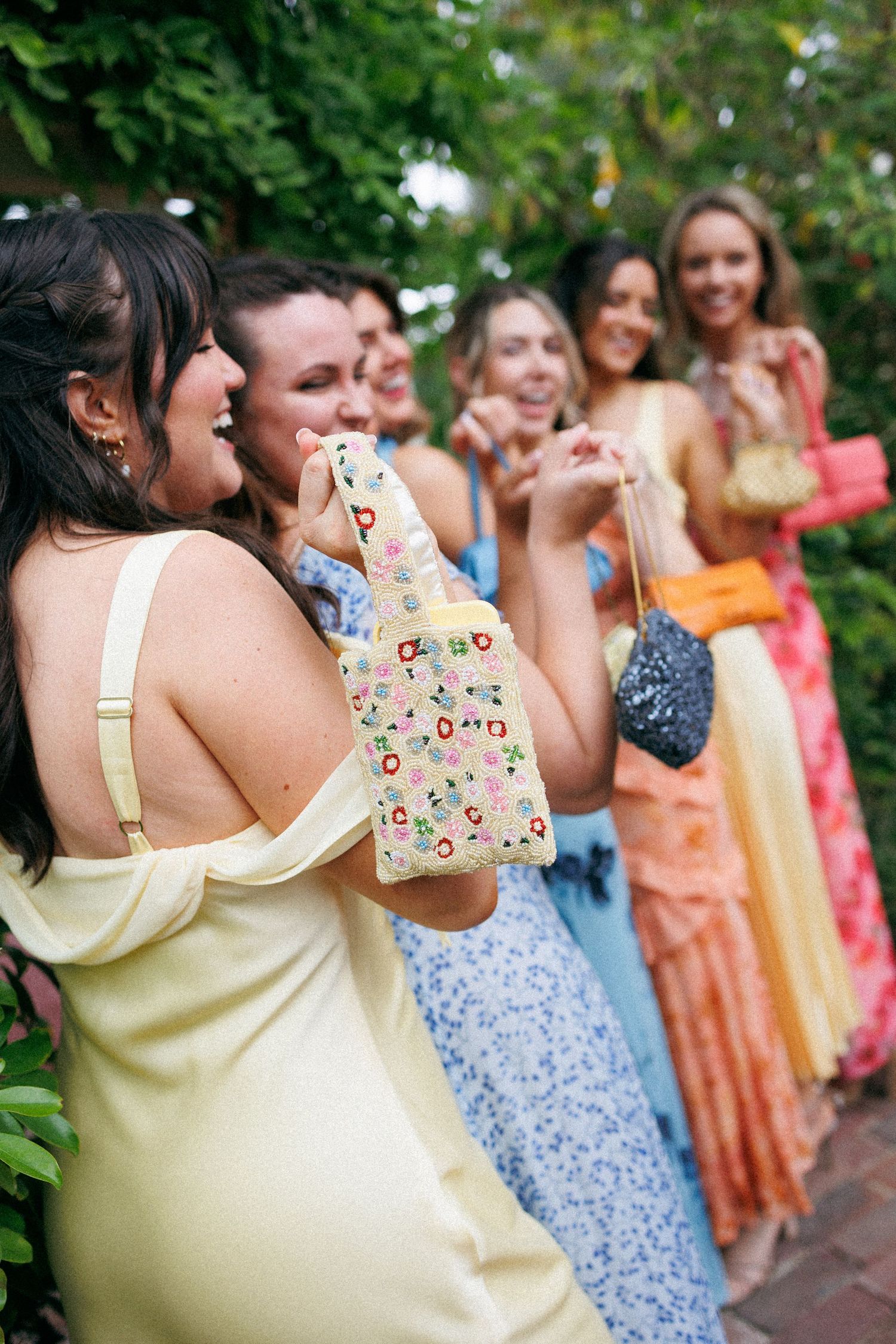 Wedding guests in colorful dresses laughing outdoors, one holding a floral water bottle.
