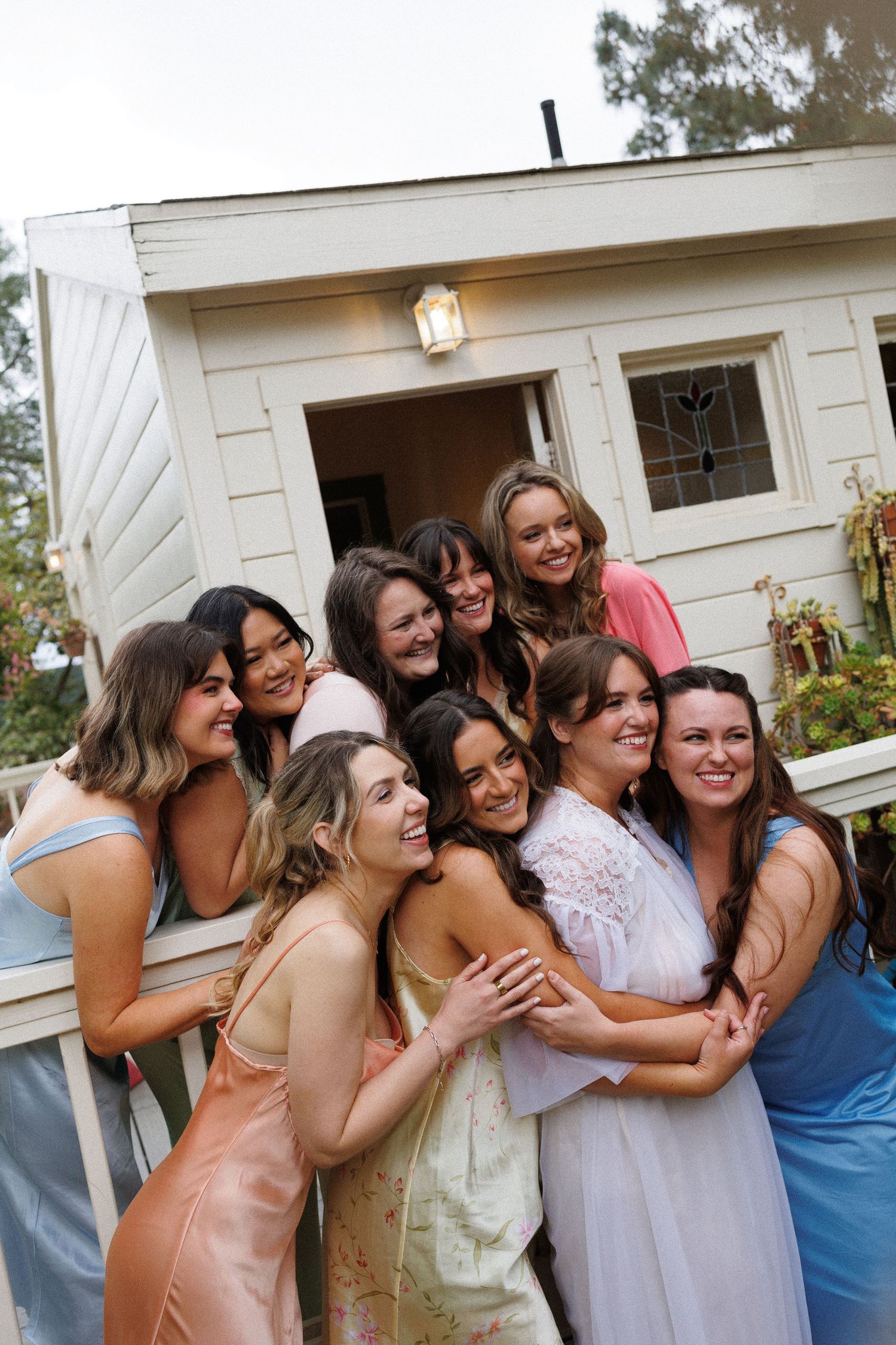 A joyful group of bridesmaids in colorful dresses hugging the bride in a white robe outside a charming white cottage.