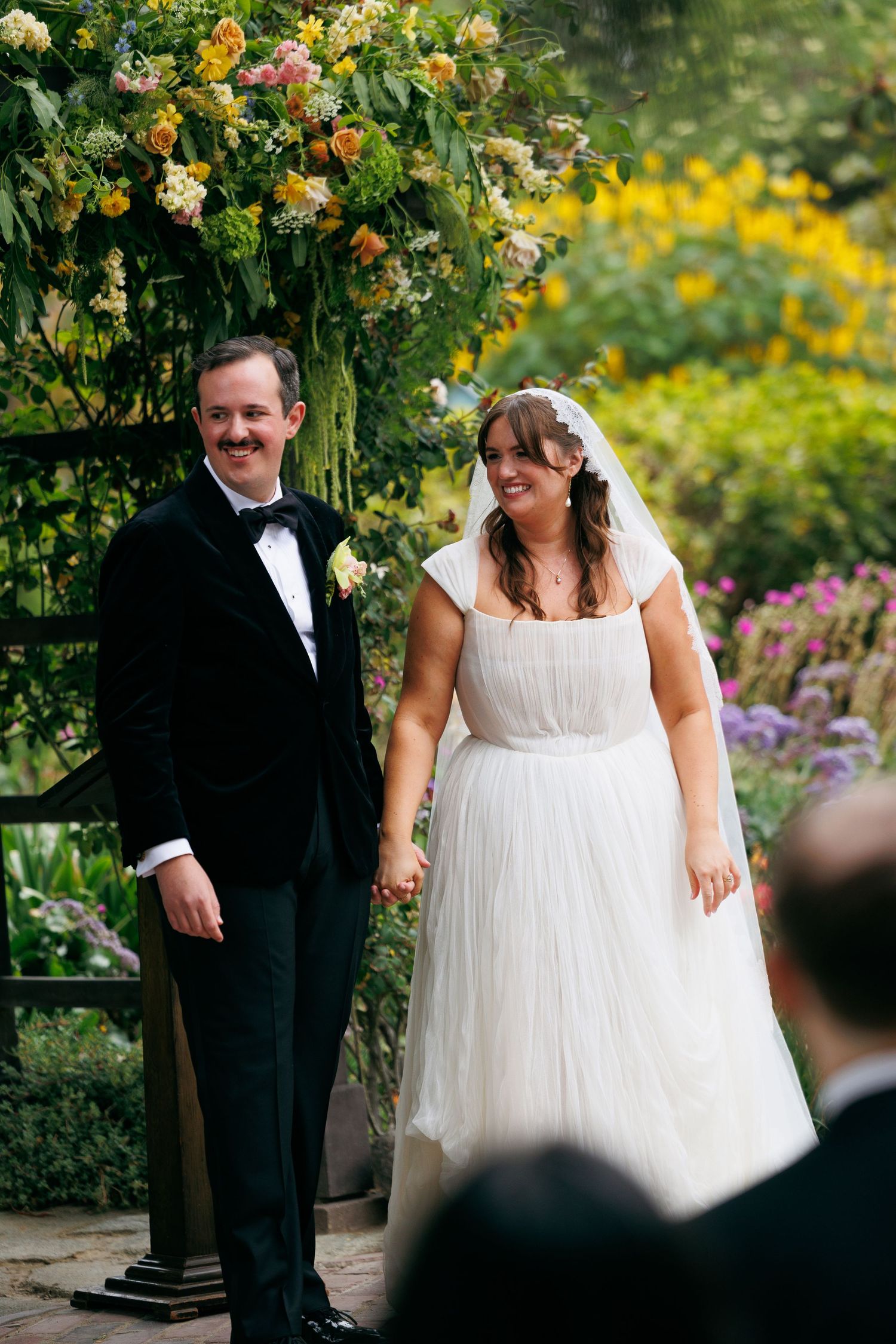 Smiling bride in white tulle gown and groom in black tuxedo stand together under floral arch in garden ceremony.