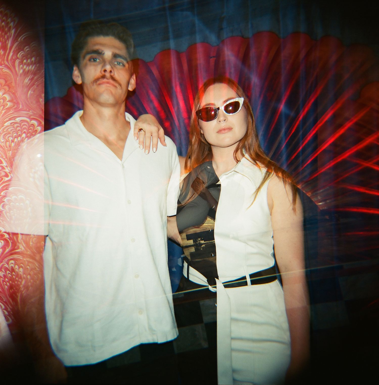 Man and woman posing together in front of red decorative backdrop, woman wearing sunglasses and white outfit.