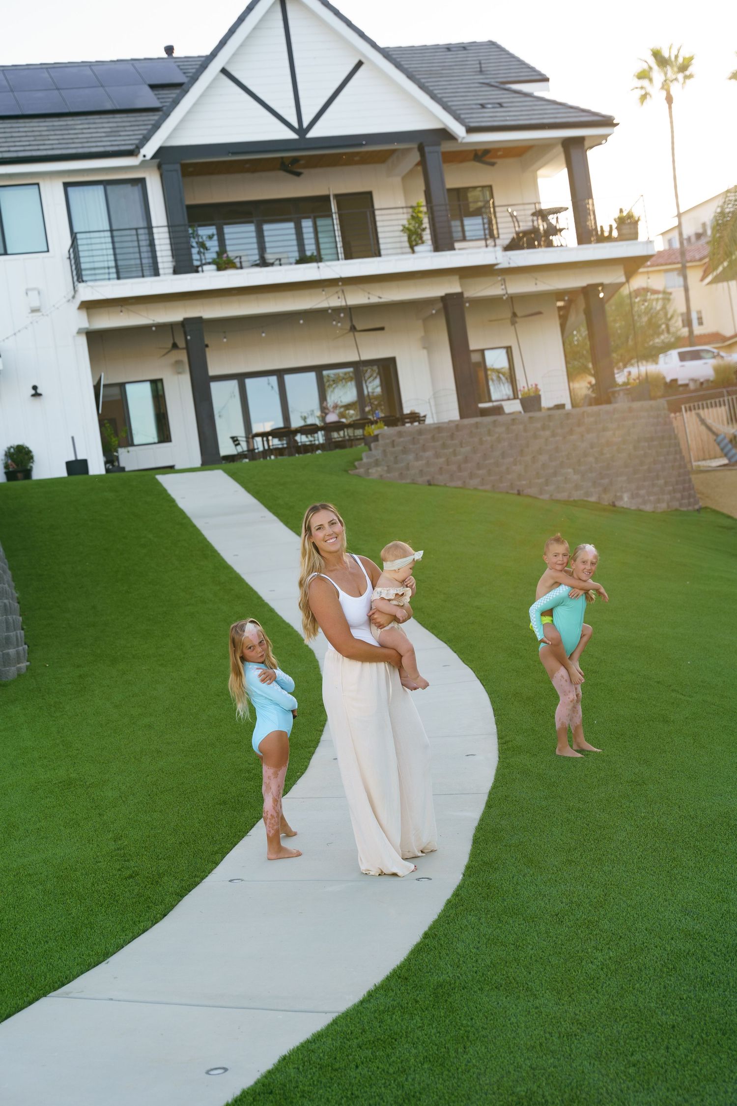 A smiling mother holds a baby on a lush lawn in front of a large modern farmhouse at sunset.