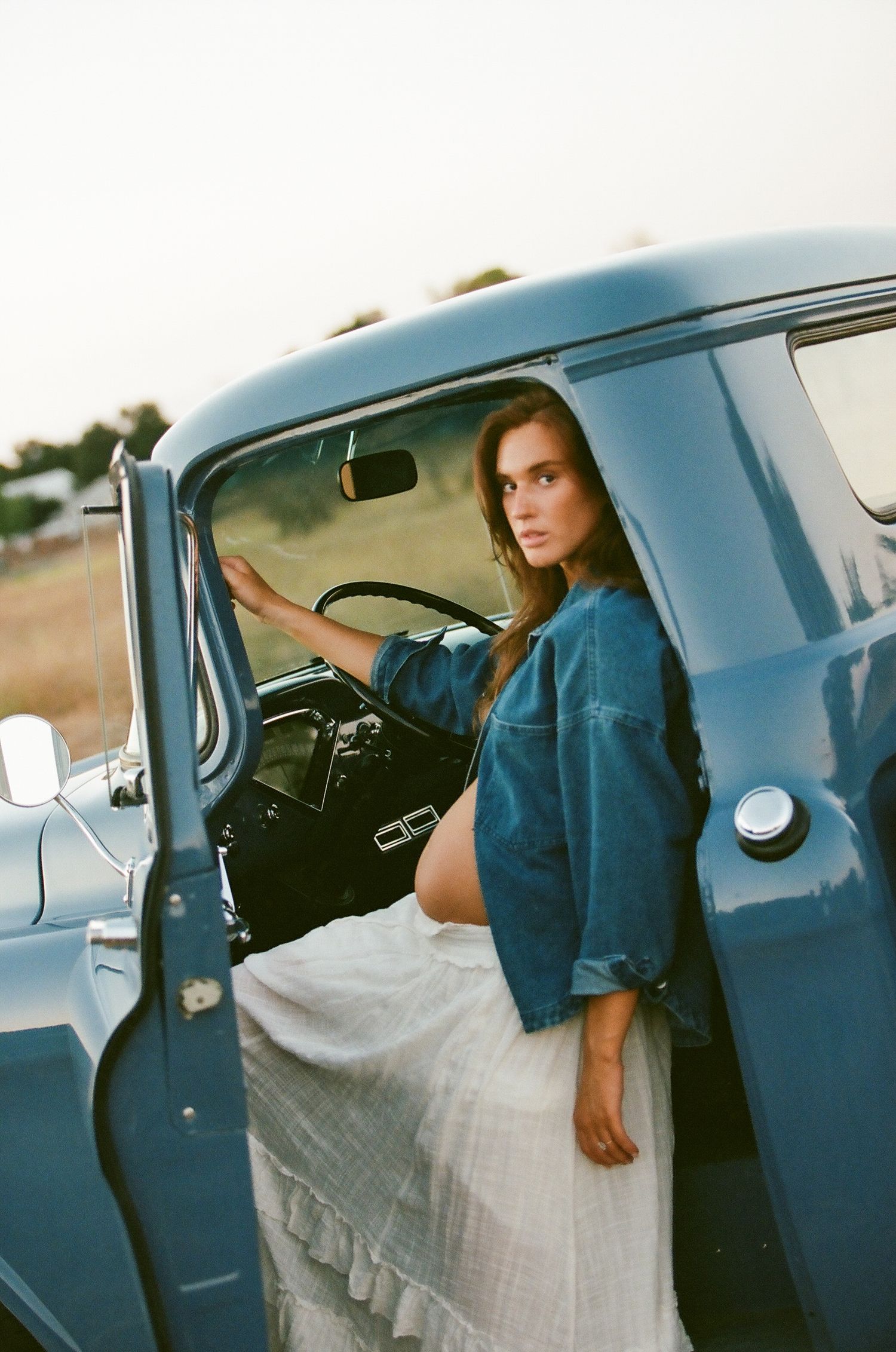 Pregnant woman in denim jacket poses in doorway of classic blue vintage truck.