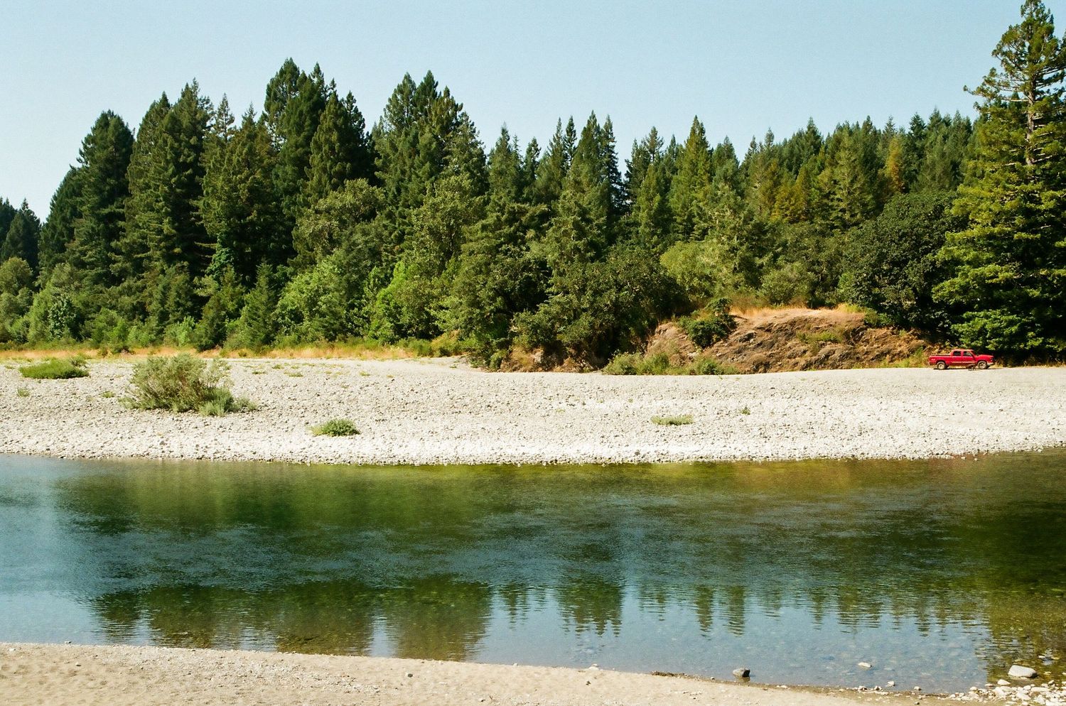 Tranquil river with rocky banks and lush evergreen forest reflected in calm clear water.