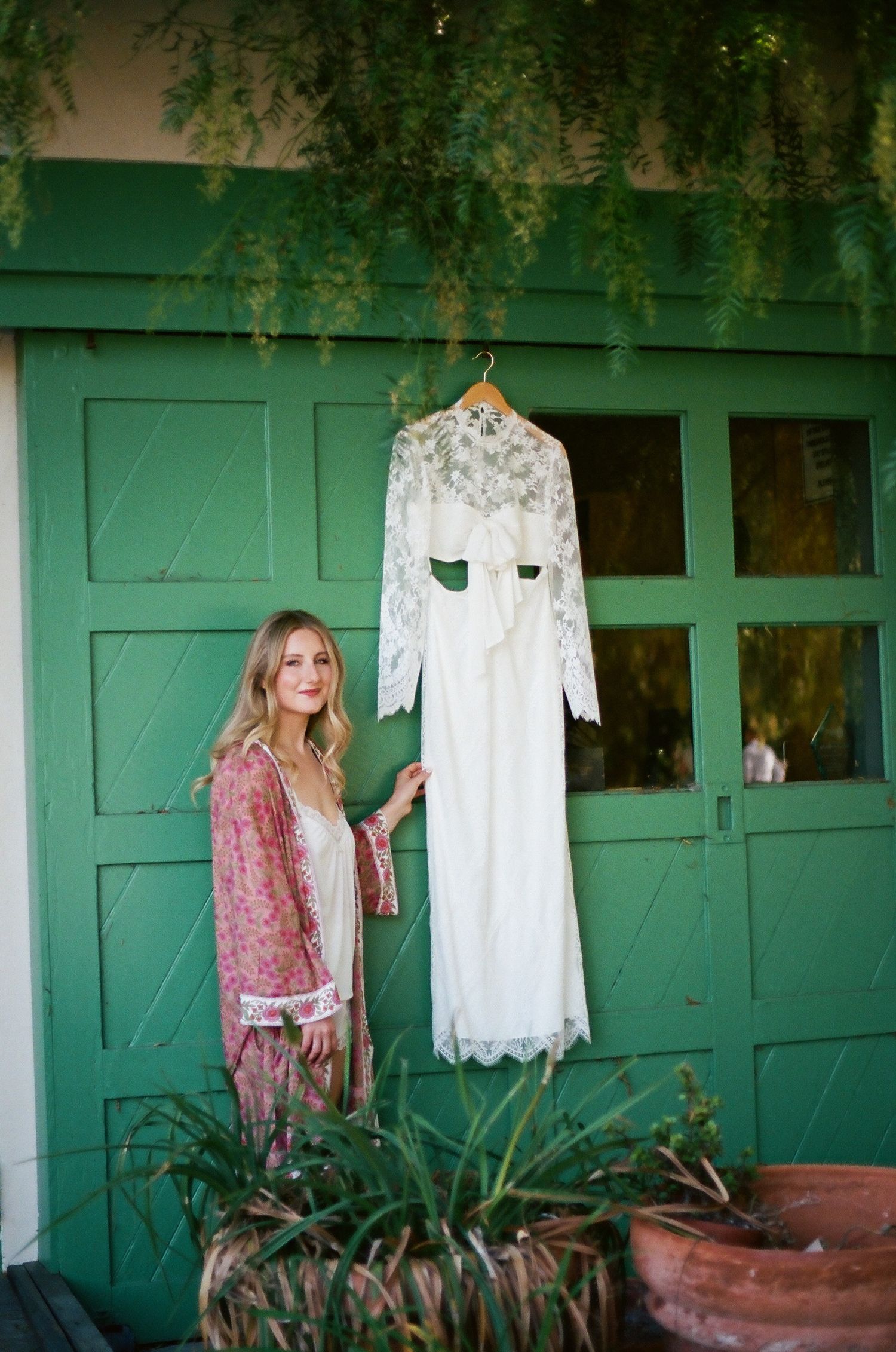 Woman in floral robe stands beside a white lace wedding dress hanging on a green door outdoors.