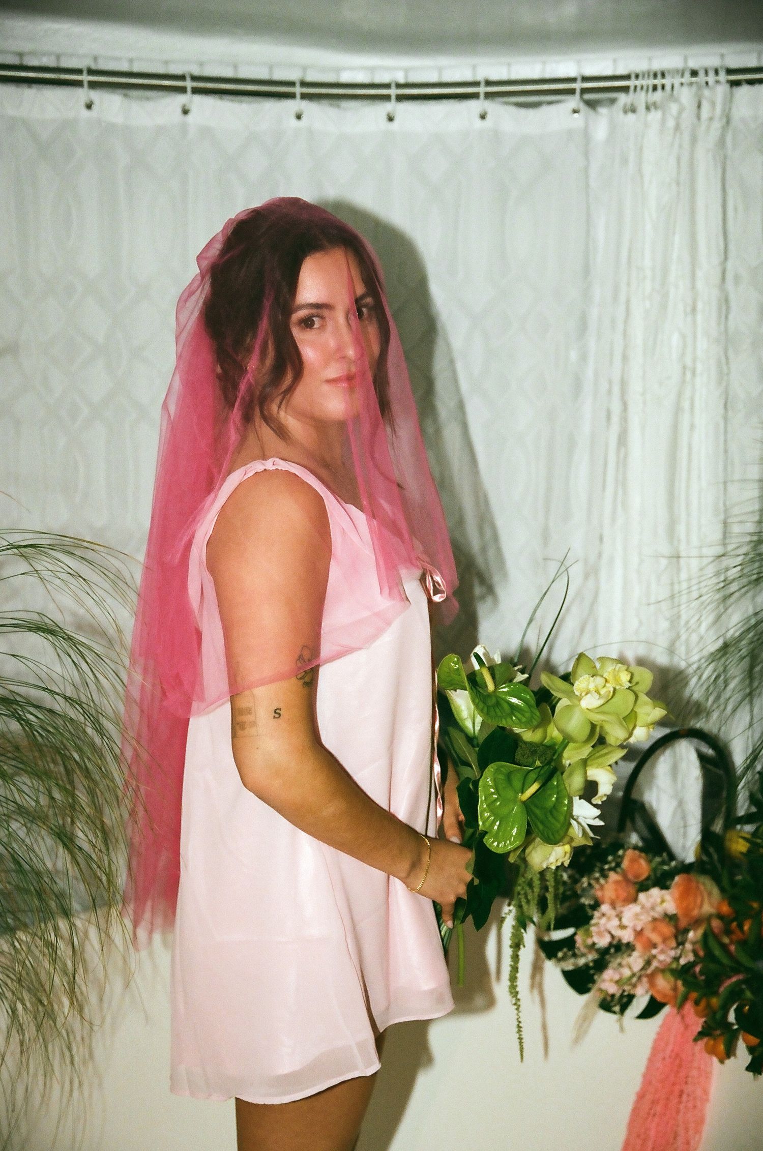 Woman in pink slip dress with pink veil holding green anthurium bouquet in bathroom setting.