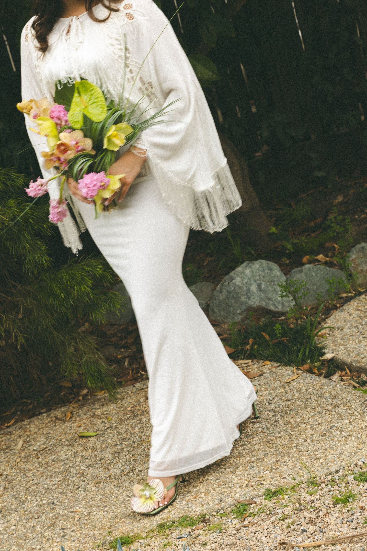 Bride in white fringe shawl and mermaid gown holding a tropical bouquet walking on a garden path.