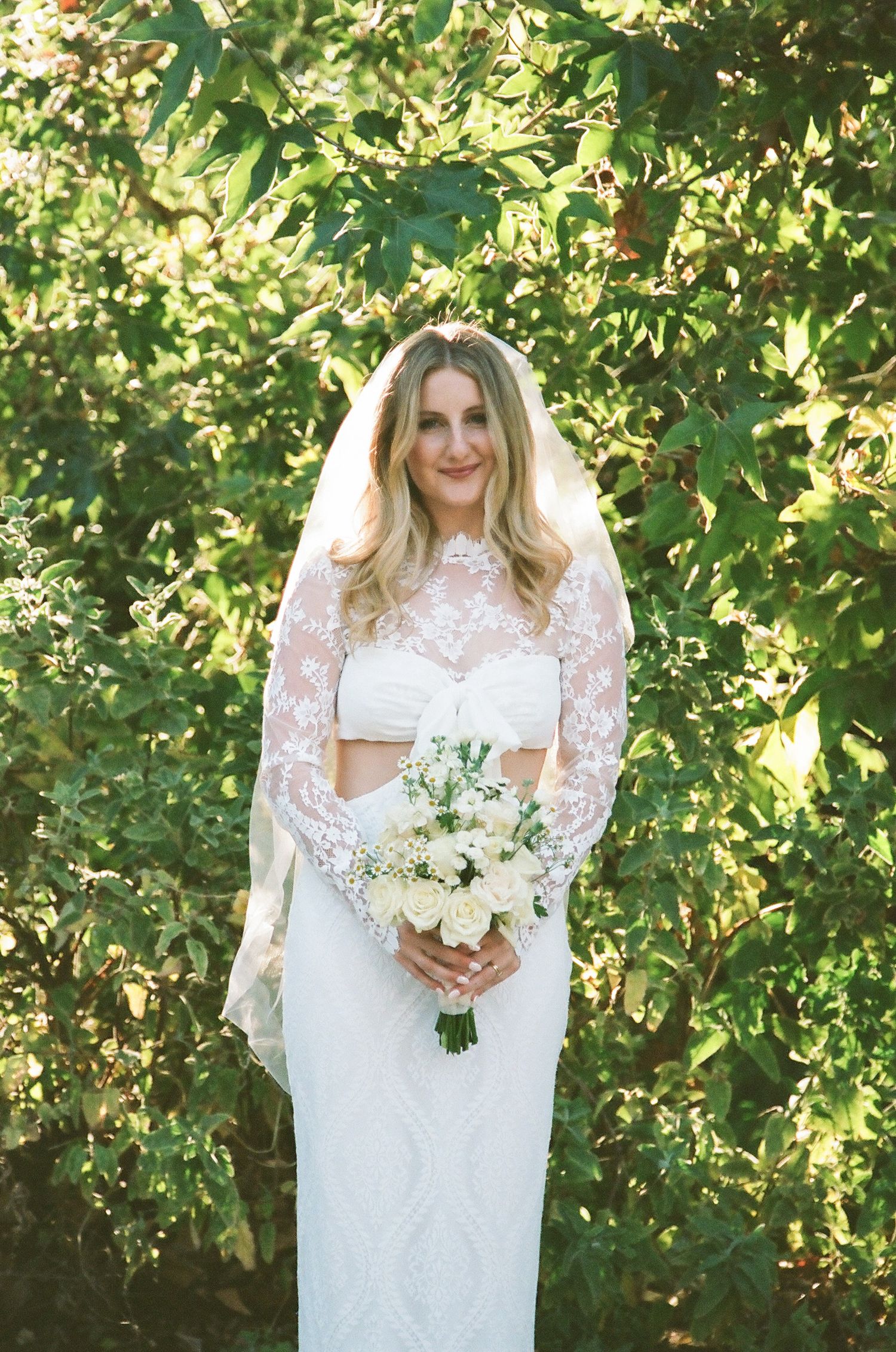 Bride in lace overlay gown and veil holds white bouquet, standing before lush green trees on wedding day.