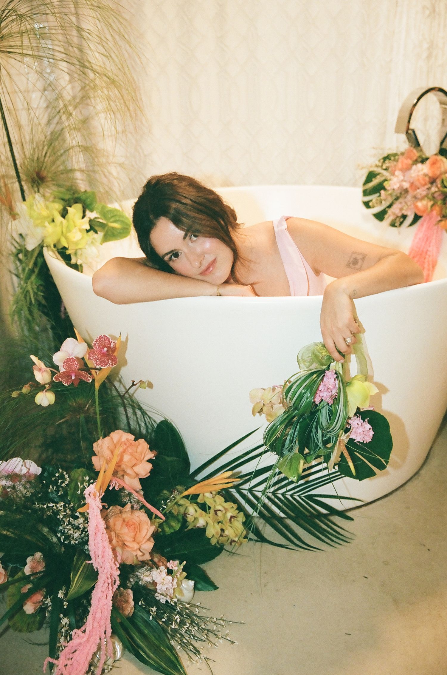 Woman in pink dress relaxing in white freestanding bathtub surrounded by tropical floral arrangements.