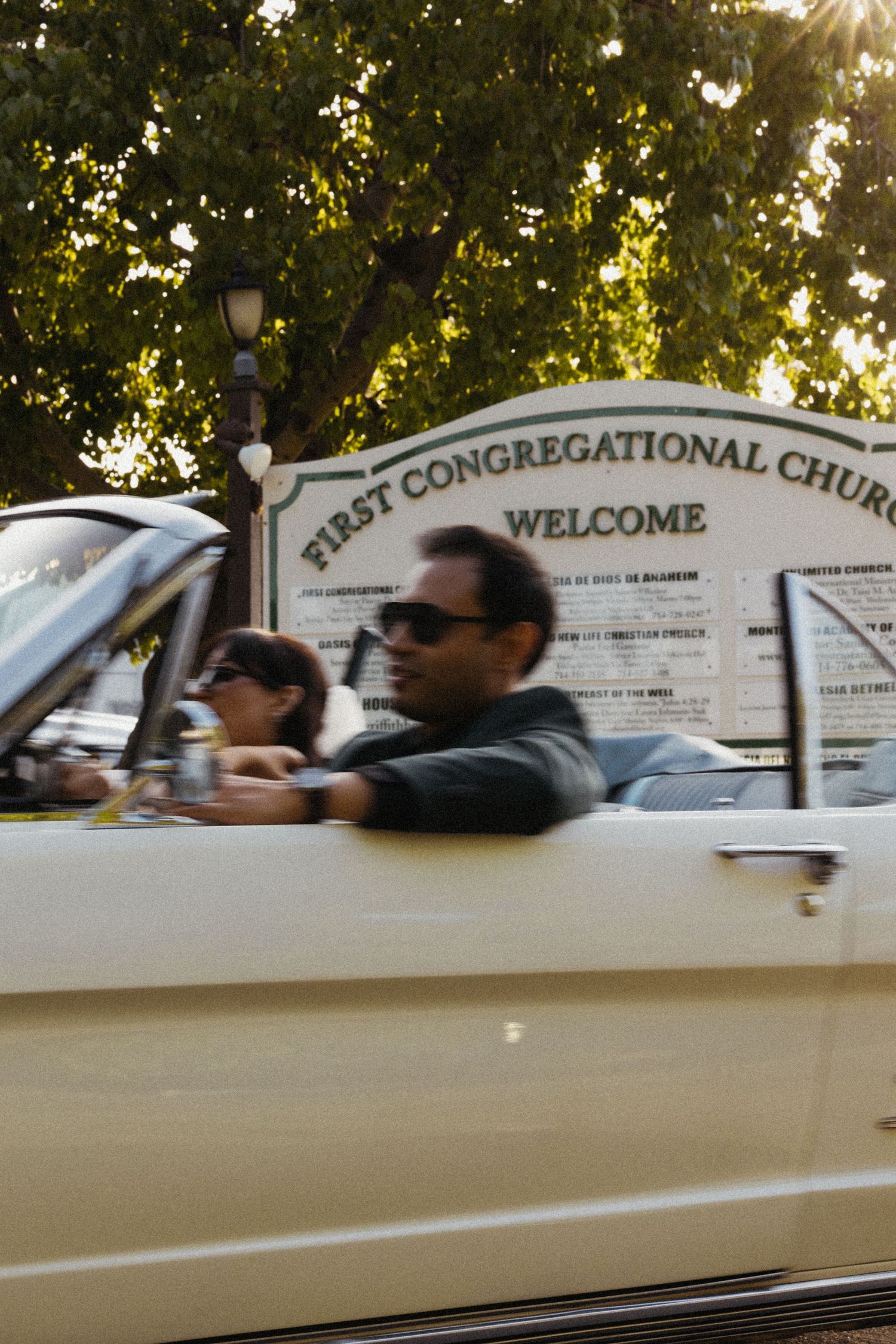 Couple in vintage convertible car outside First Congregational Church welcome sign on sunny day.