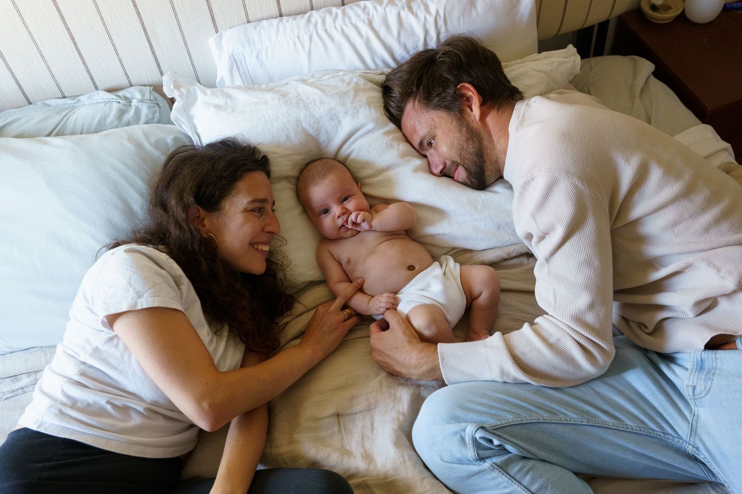 Two parents smile at their laughing baby lying between them on a cozy white bed.