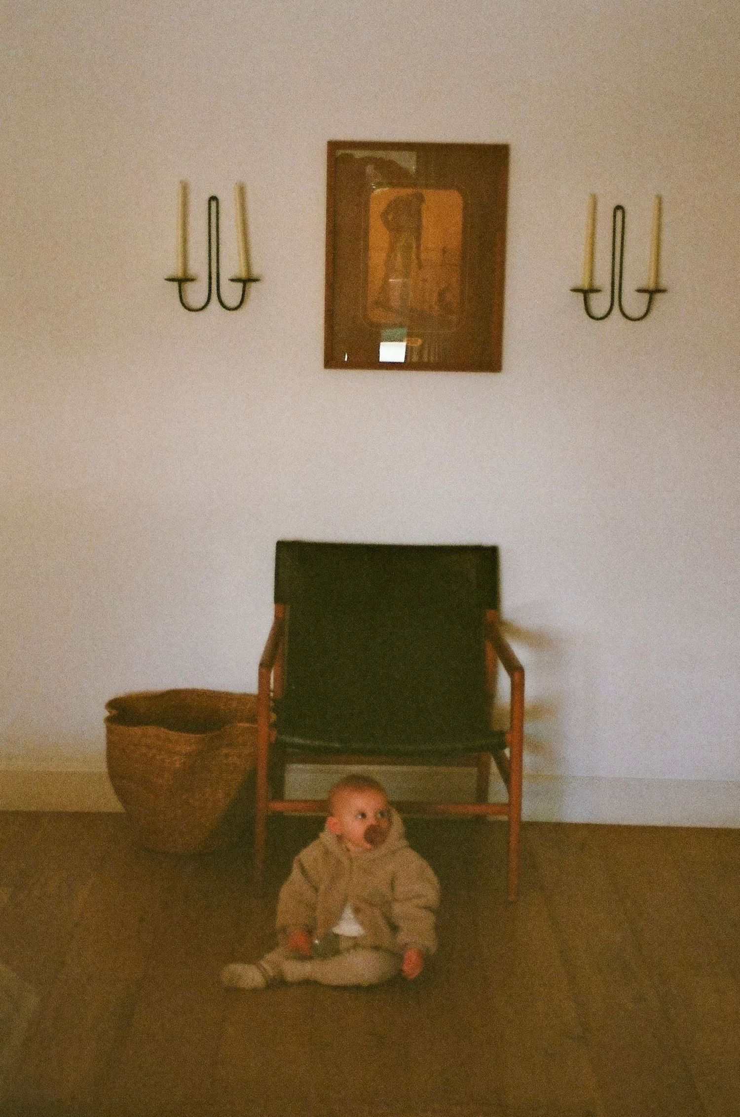 Baby in beige outfit sits on wood floor beside green chair flanked by brass wall sconces.