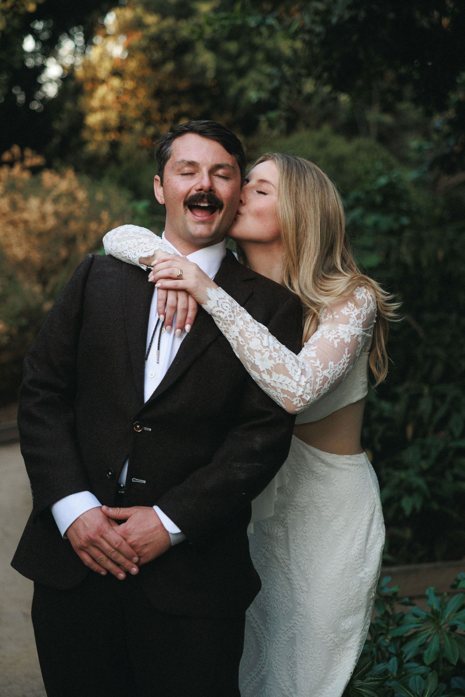 Bride in lace gown kisses groom in dark suit outdoors at garden wedding ceremony.
