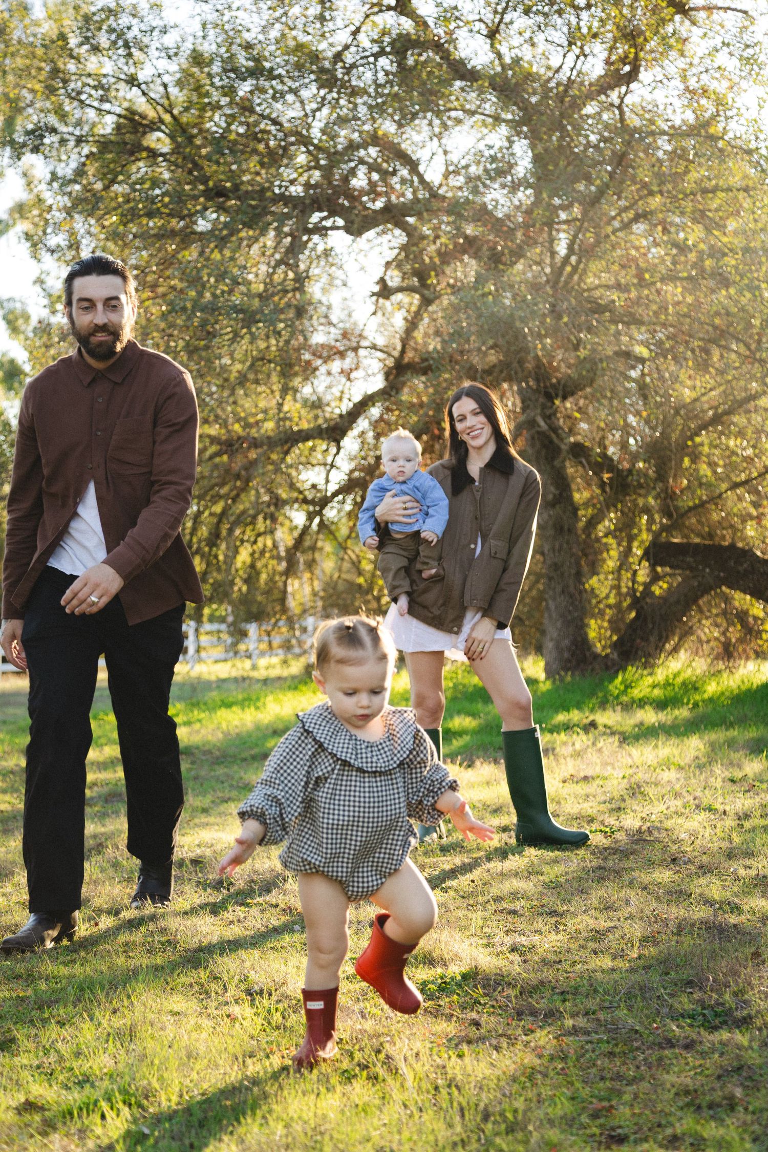 Family of four enjoying sunny park, dad in brown shirt, mom holding baby in green boots, toddler girl running in plaid dress.