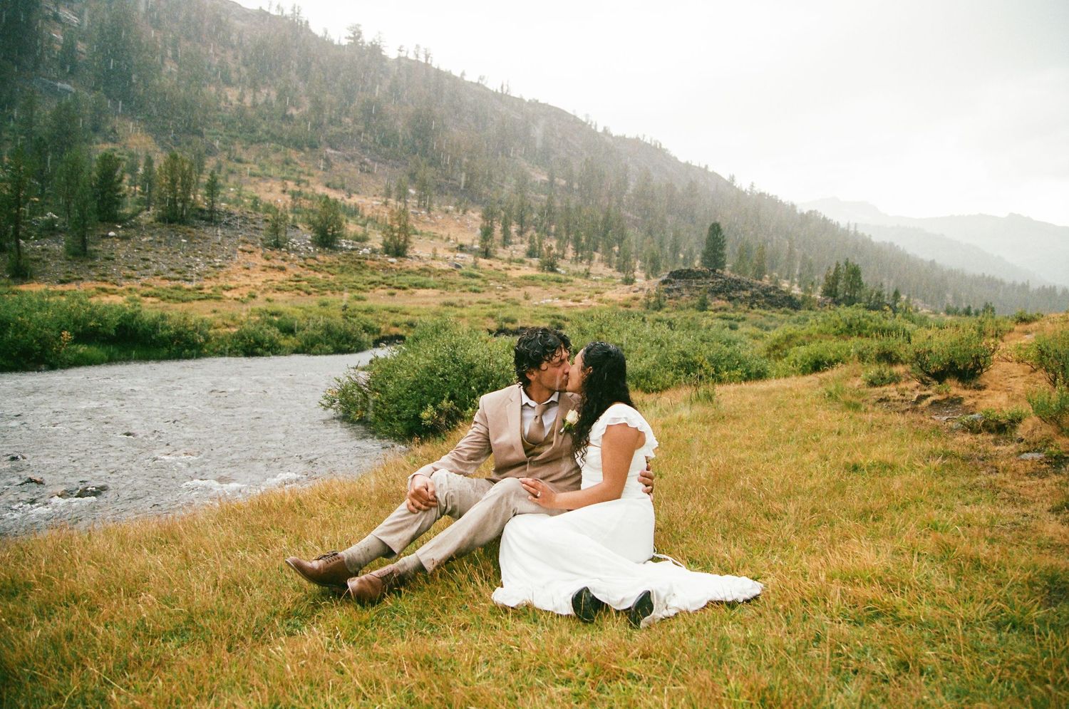 Couple kissing while sitting on grass by a mountain river on their wedding day in a scenic forest setting.