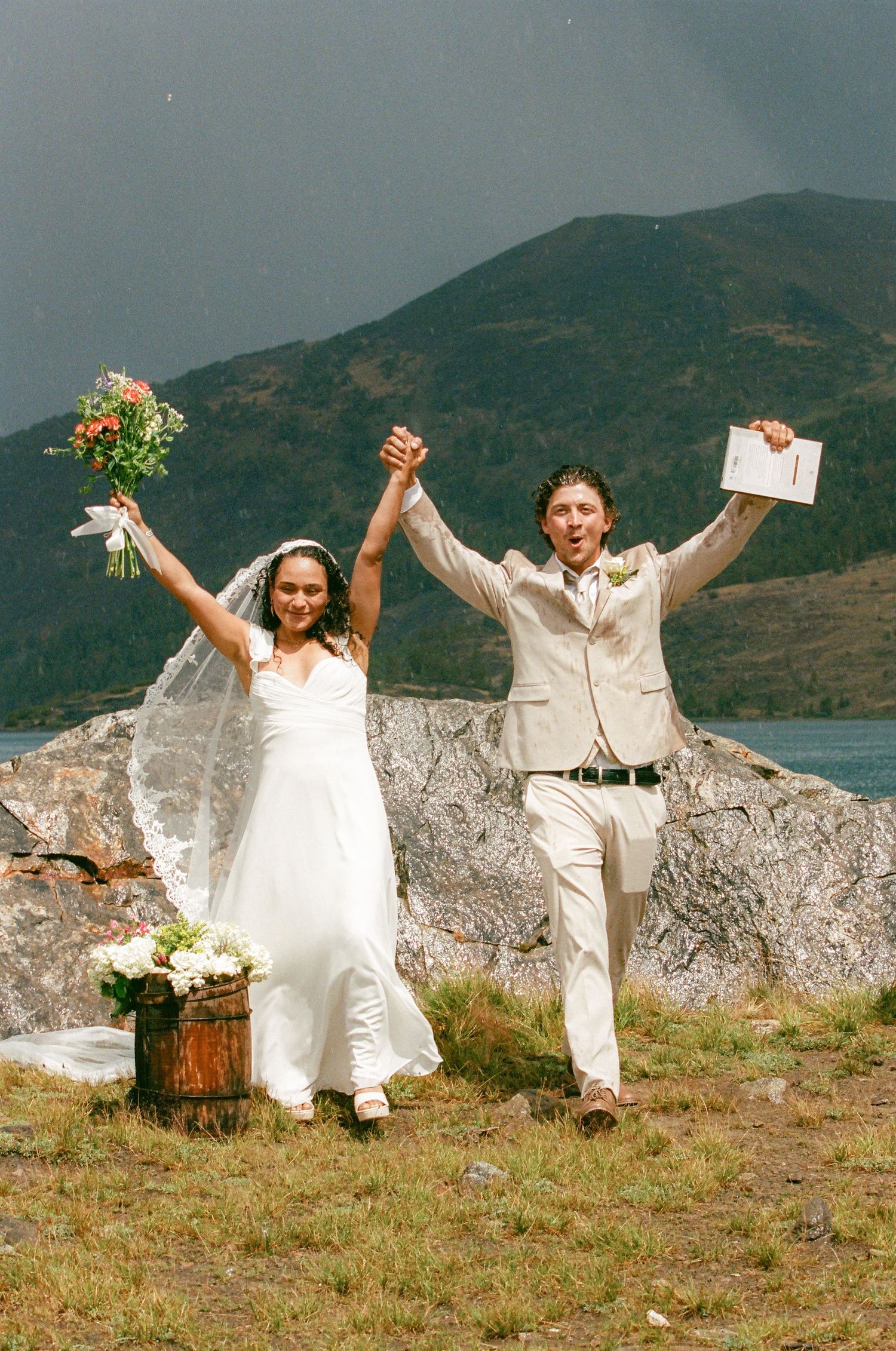 Joyful newlywed couple celebrating outdoors with mountains and lake backdrop, bride in white gown holding bouquet, groom in tan suit.