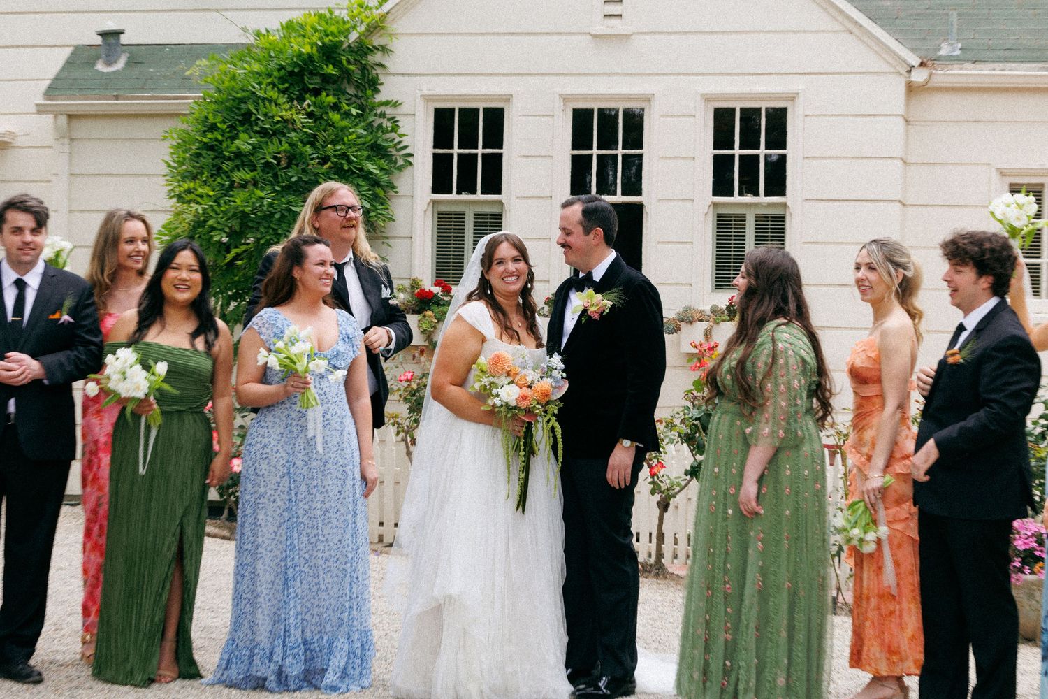 Wedding party gathered outside an elegant venue with bridesmaids in green and floral dresses.