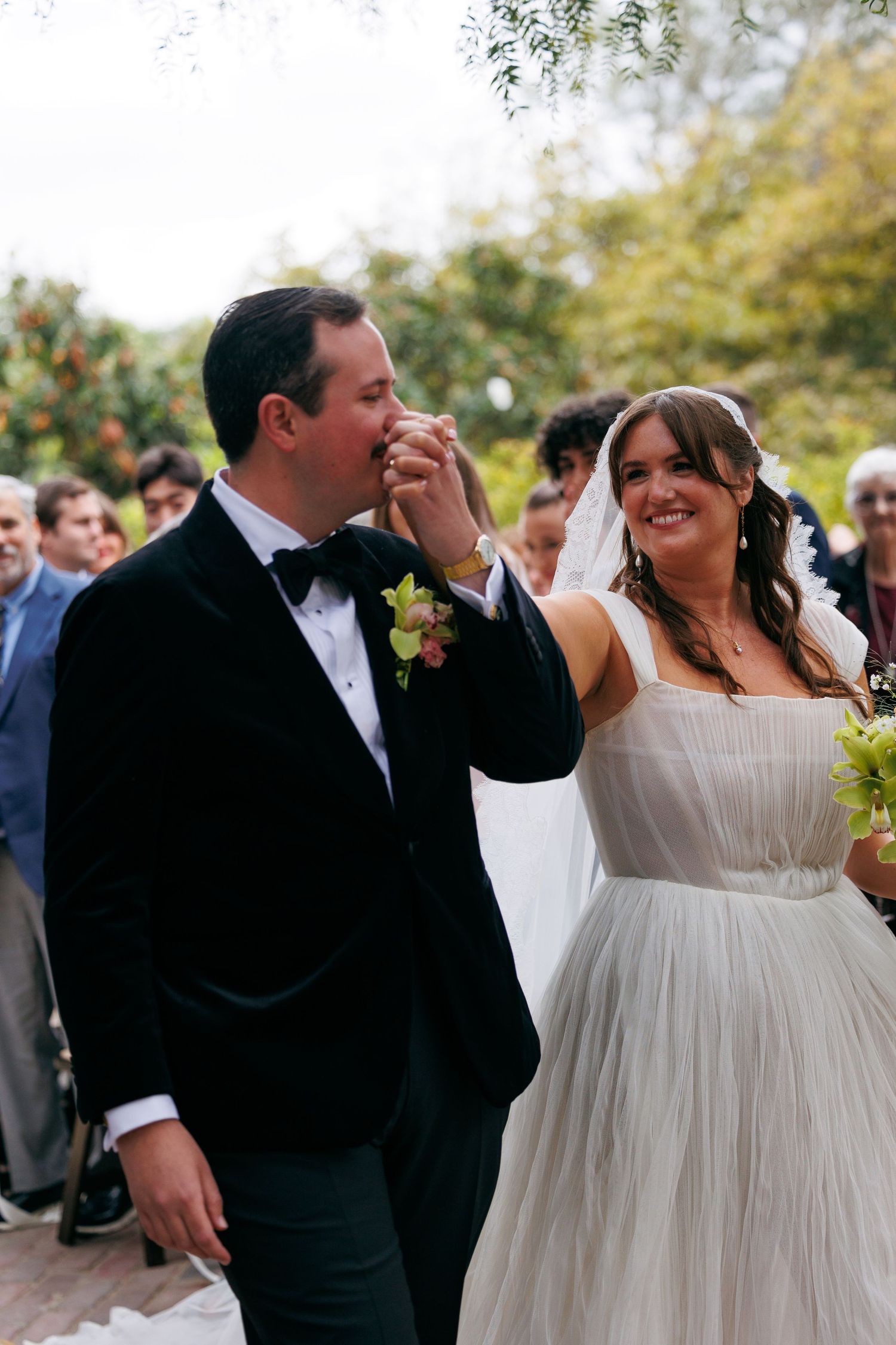 Groom in black tuxedo kisses bride's hand as she smiles in white tulle gown after outdoor garden wedding ceremony.