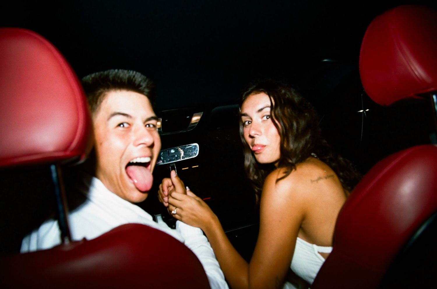 Playful couple laughing in a car at night with red leather seats in the background.