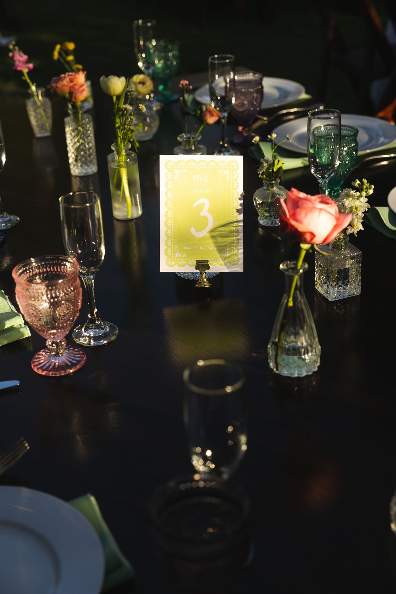 Dark romantic wedding reception table with colorful vintage goblets, bud vases with flowers, and illuminated table number 3.