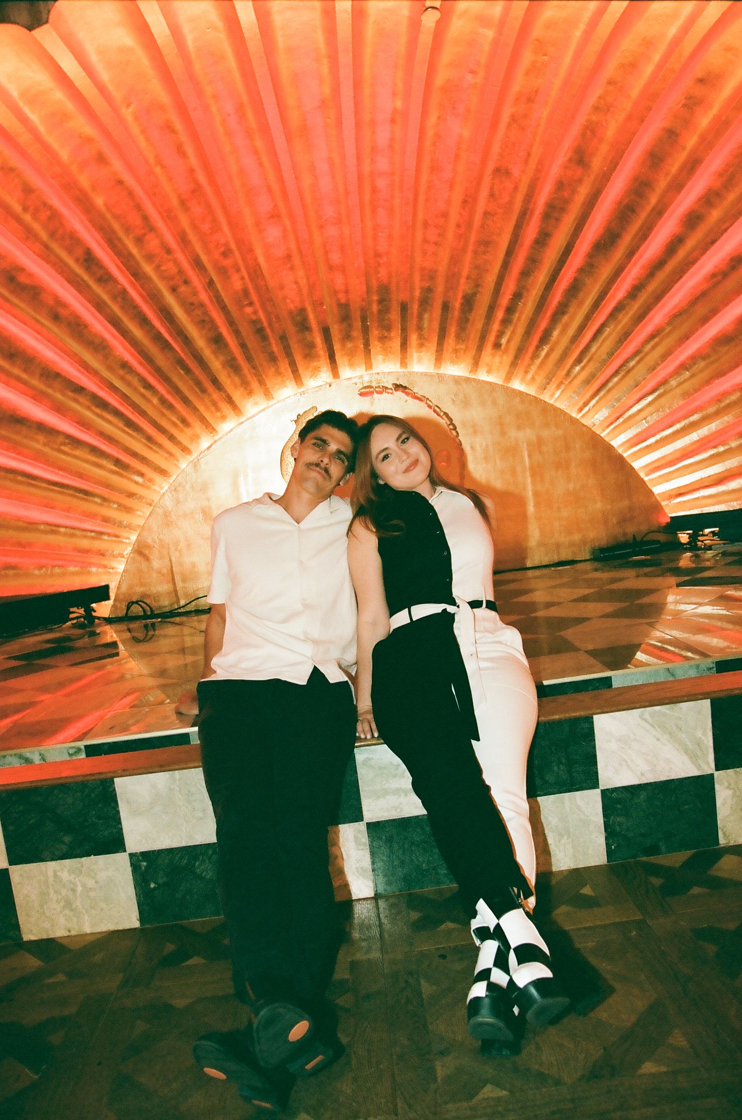 Couple sitting together on a checkered floor beneath a dramatic red sunburst arch.