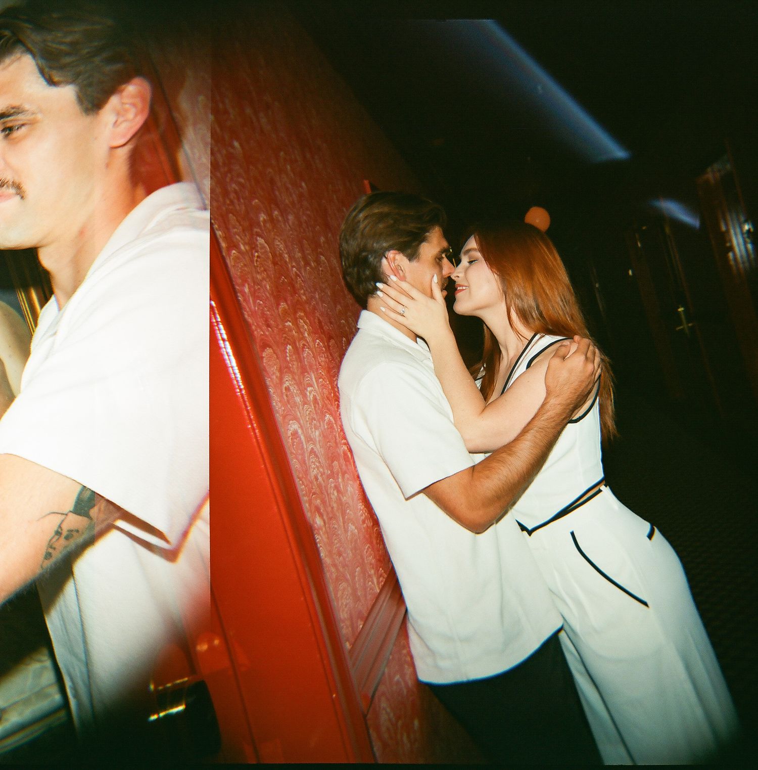 A couple shares a romantic kiss in a dimly lit red interior, captured in a film photography style.