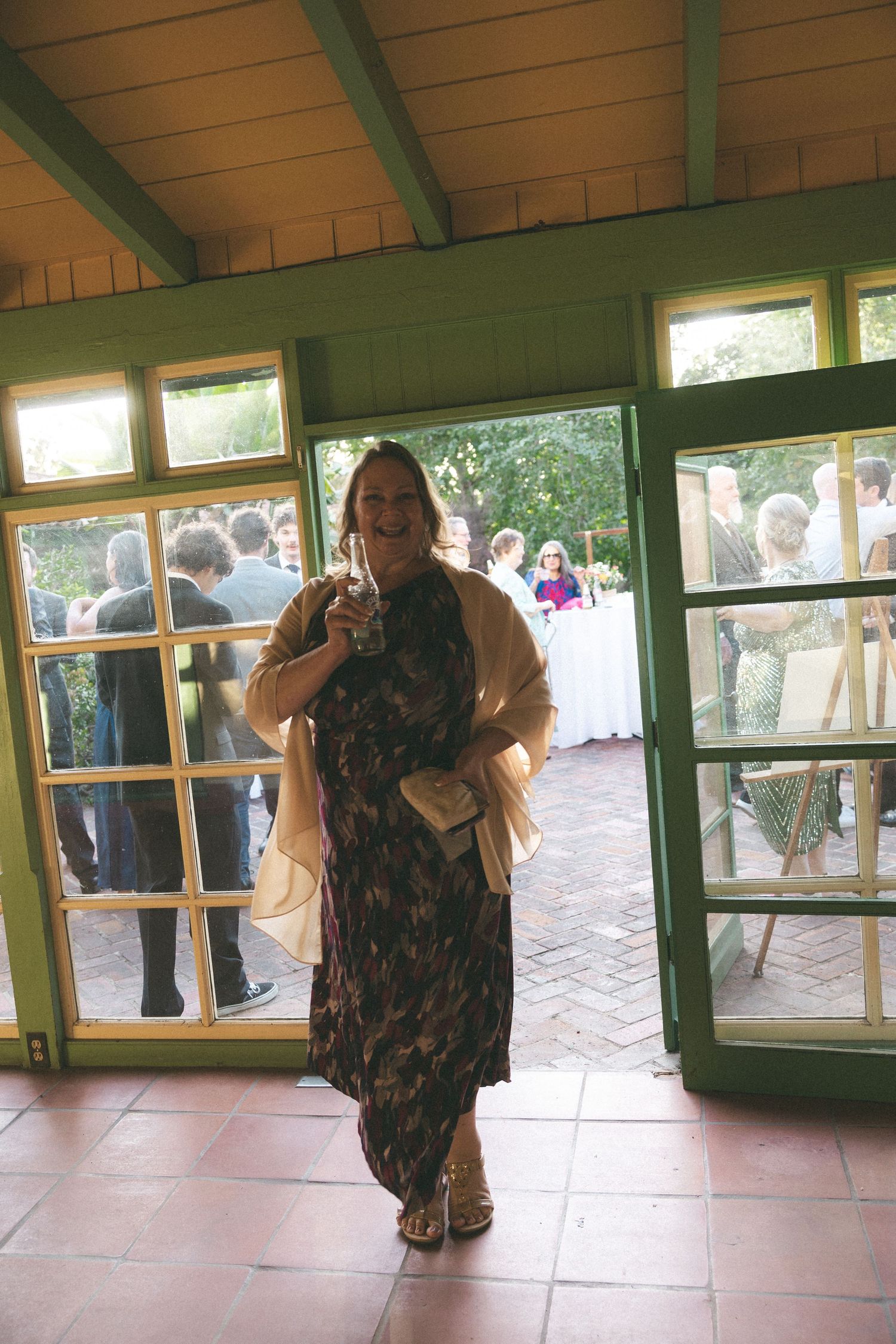 Woman in floral dress holding drink smiles at camera in green pavilion doorway with wedding guests gathered outside.