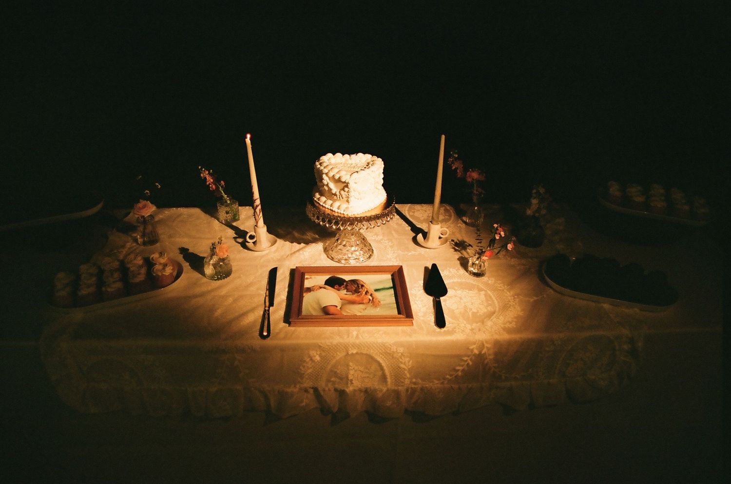 Elegant wedding cake table with white frosted cake, candles, and framed photo displayed under warm romantic lighting.