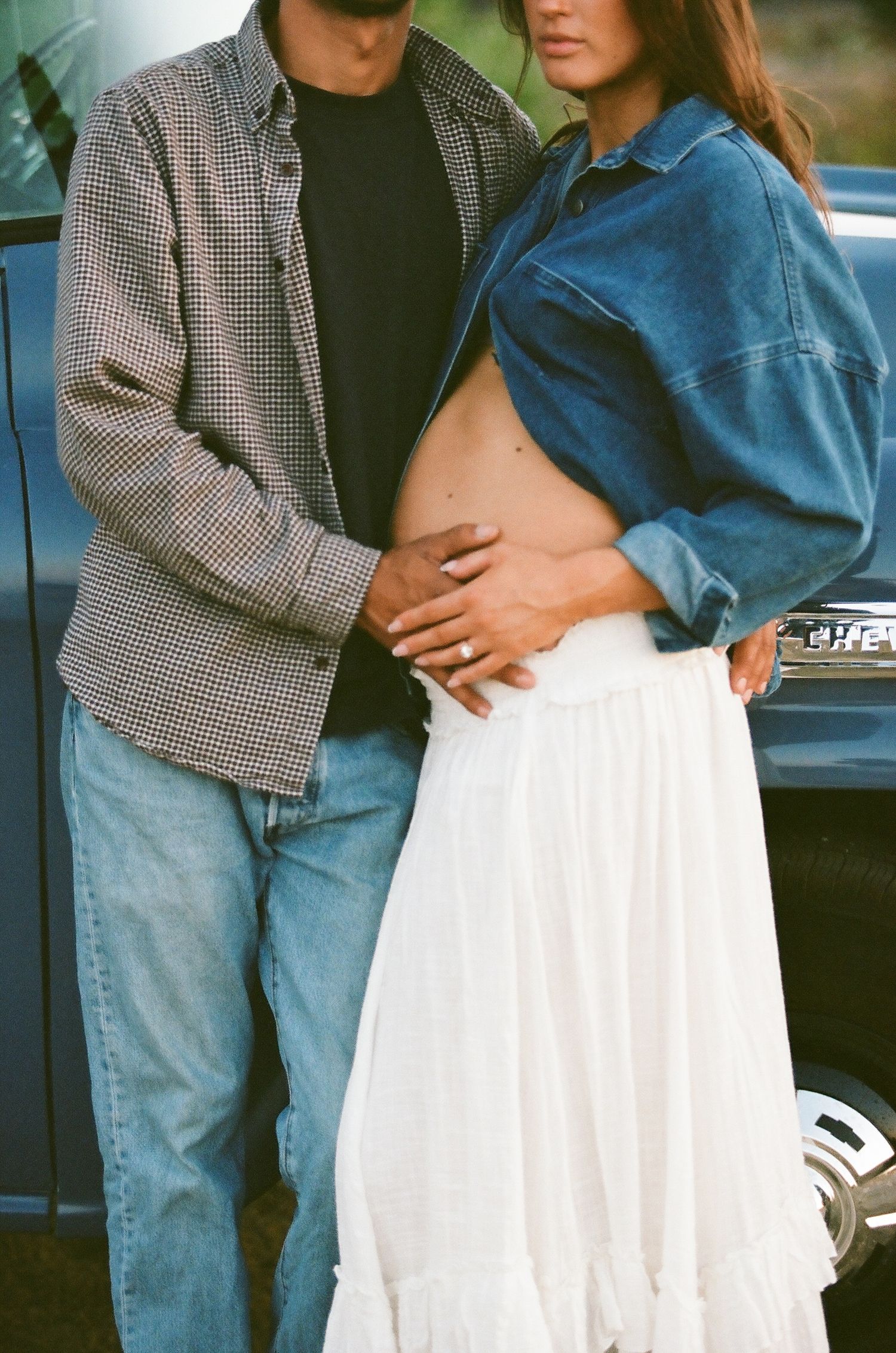 Couple sharing an intimate maternity moment outdoors, woman in denim jacket and white skirt revealing baby bump.