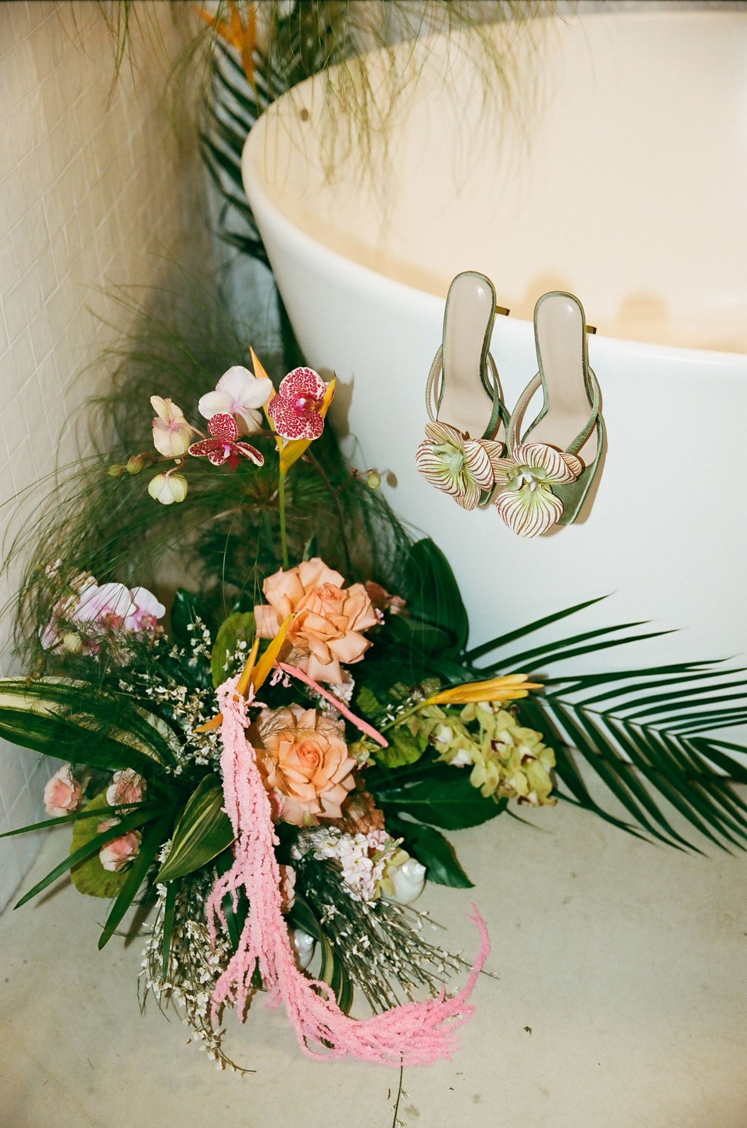 Tropical wedding bouquet with peach roses and orchids beside floral heels near a white bathtub.