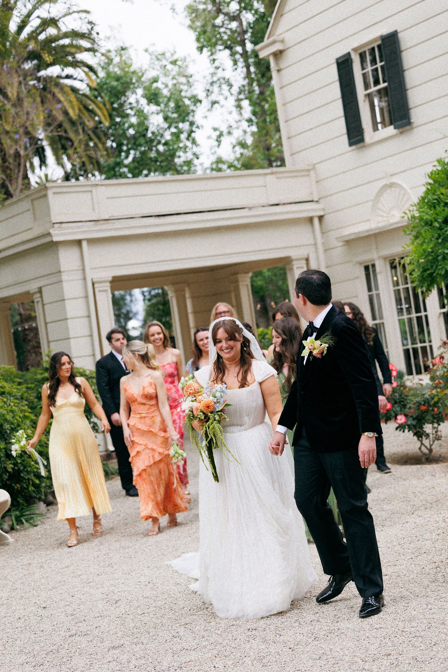 Bride in white gown and groom in black tux walk hand-in-hand with wedding party following outside elegant white estate.