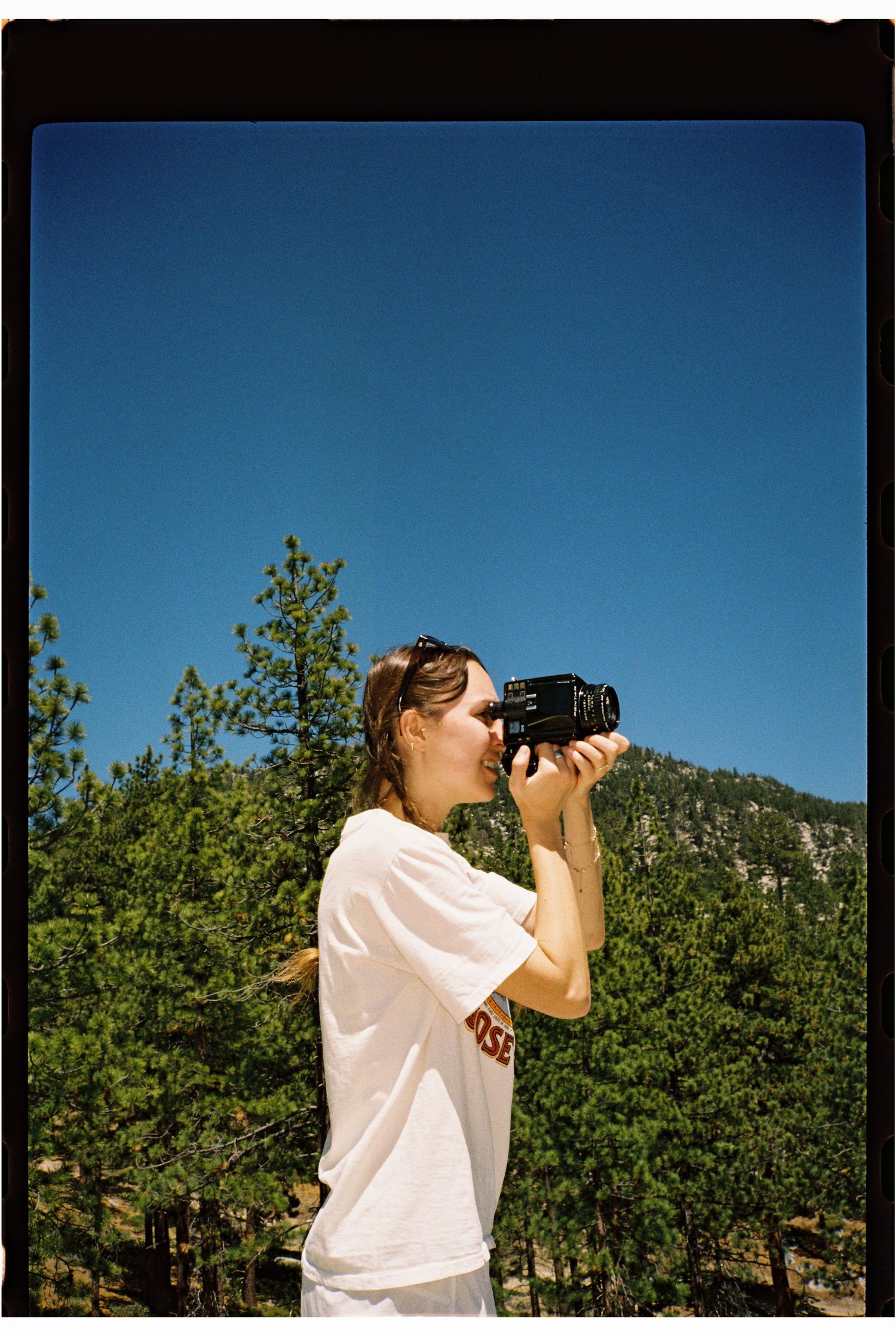 A woman in a white t-shirt photographs scenery with a camera outdoors, surrounded by pine trees under a clear blue sky.