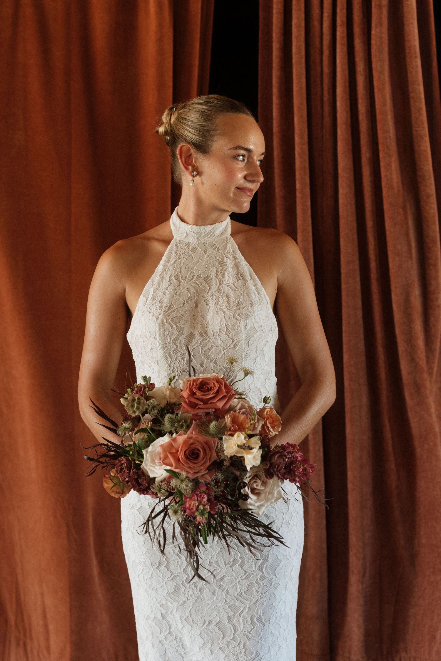 Bride in white halter lace gown holding autumn floral bouquet with roses, standing before rust-colored velvet curtains.