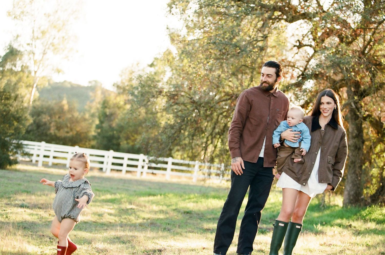 Family of four enjoying a sunny outdoor walk in a green meadow with white fence and oak trees in background.