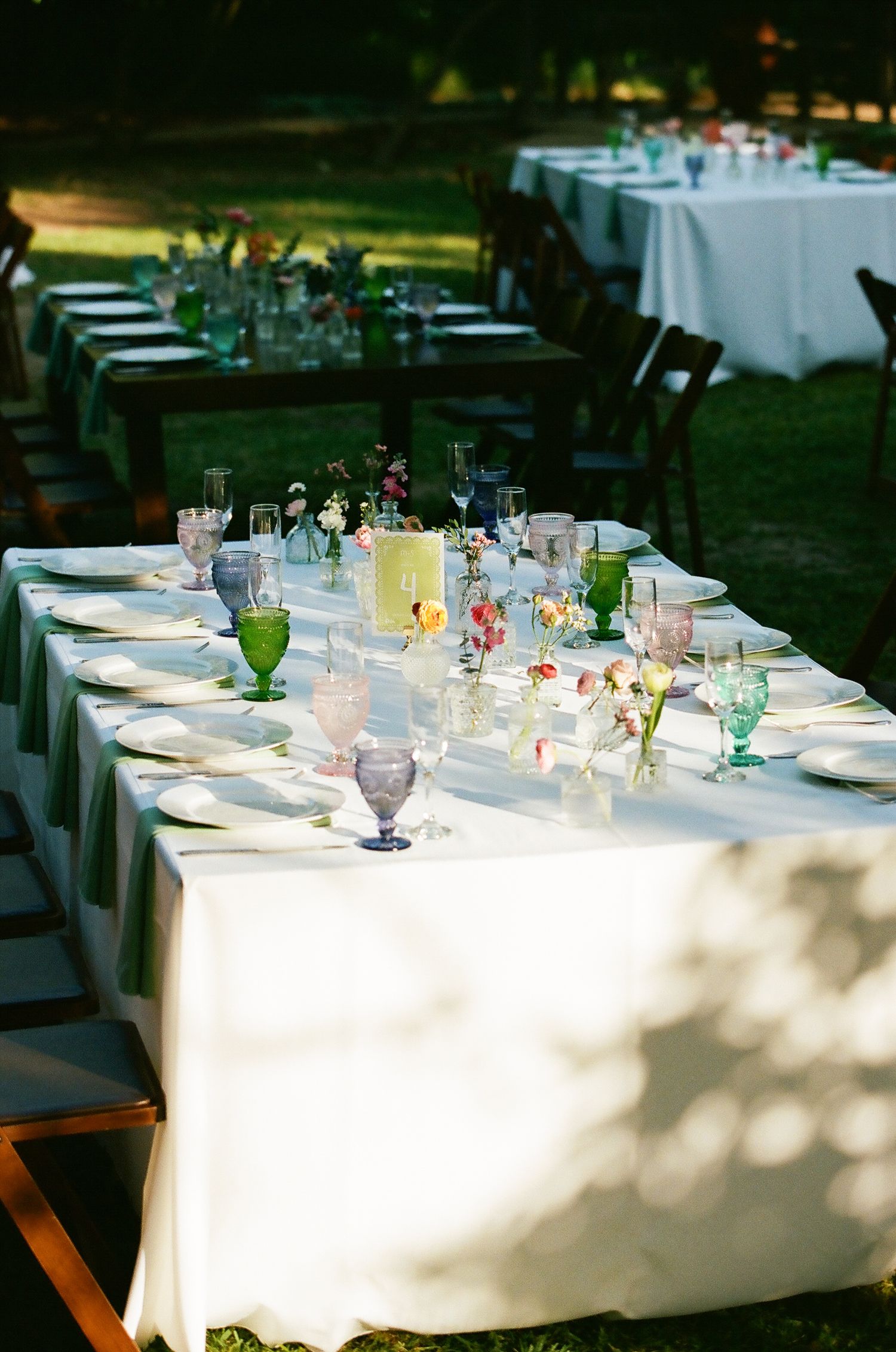 Elegant outdoor garden wedding reception with long white tablecloths, colorful glassware, and floral centerpieces in natural light.