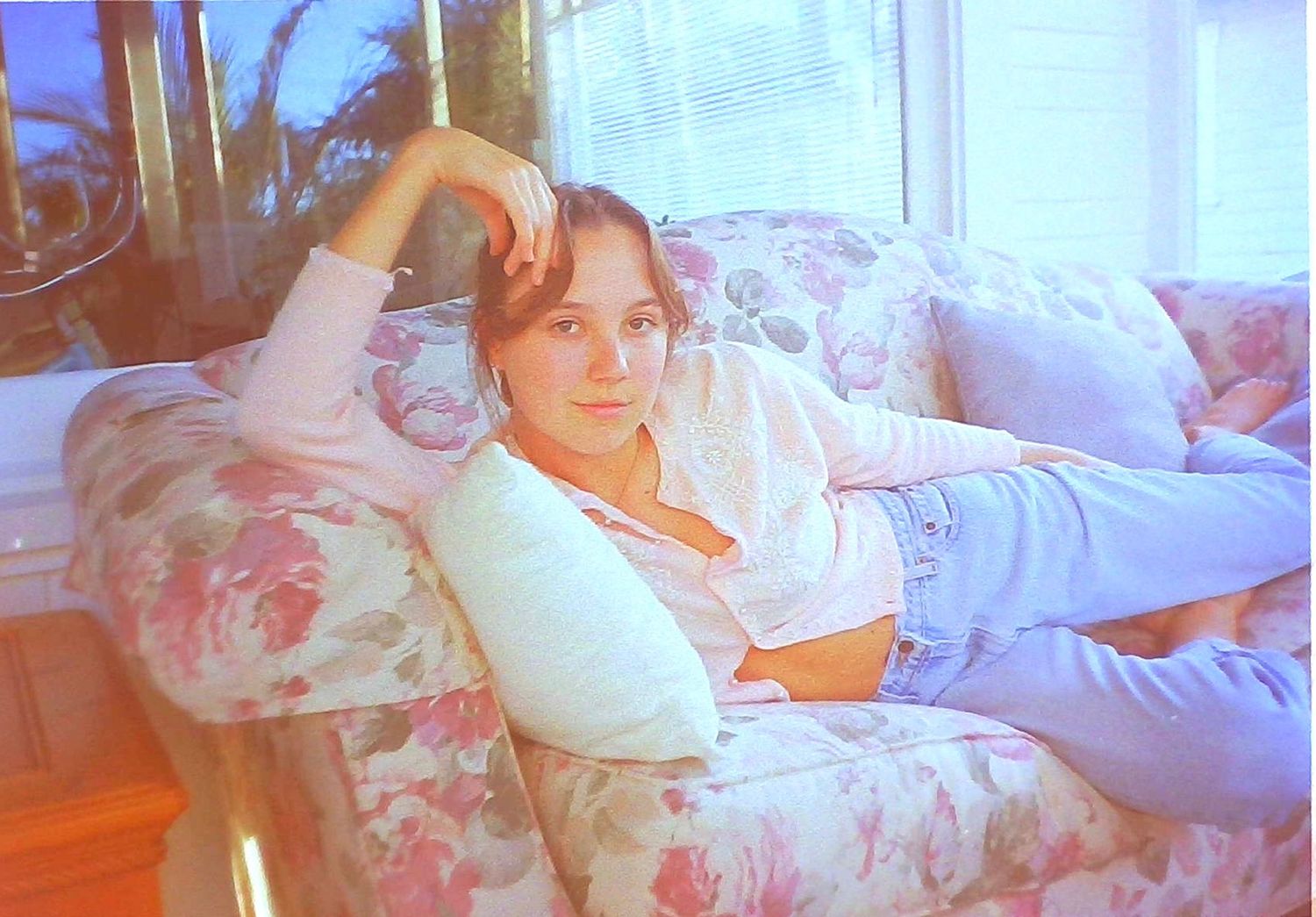 Young woman relaxing on a floral couch by a sunny window in a vintage film photo.
