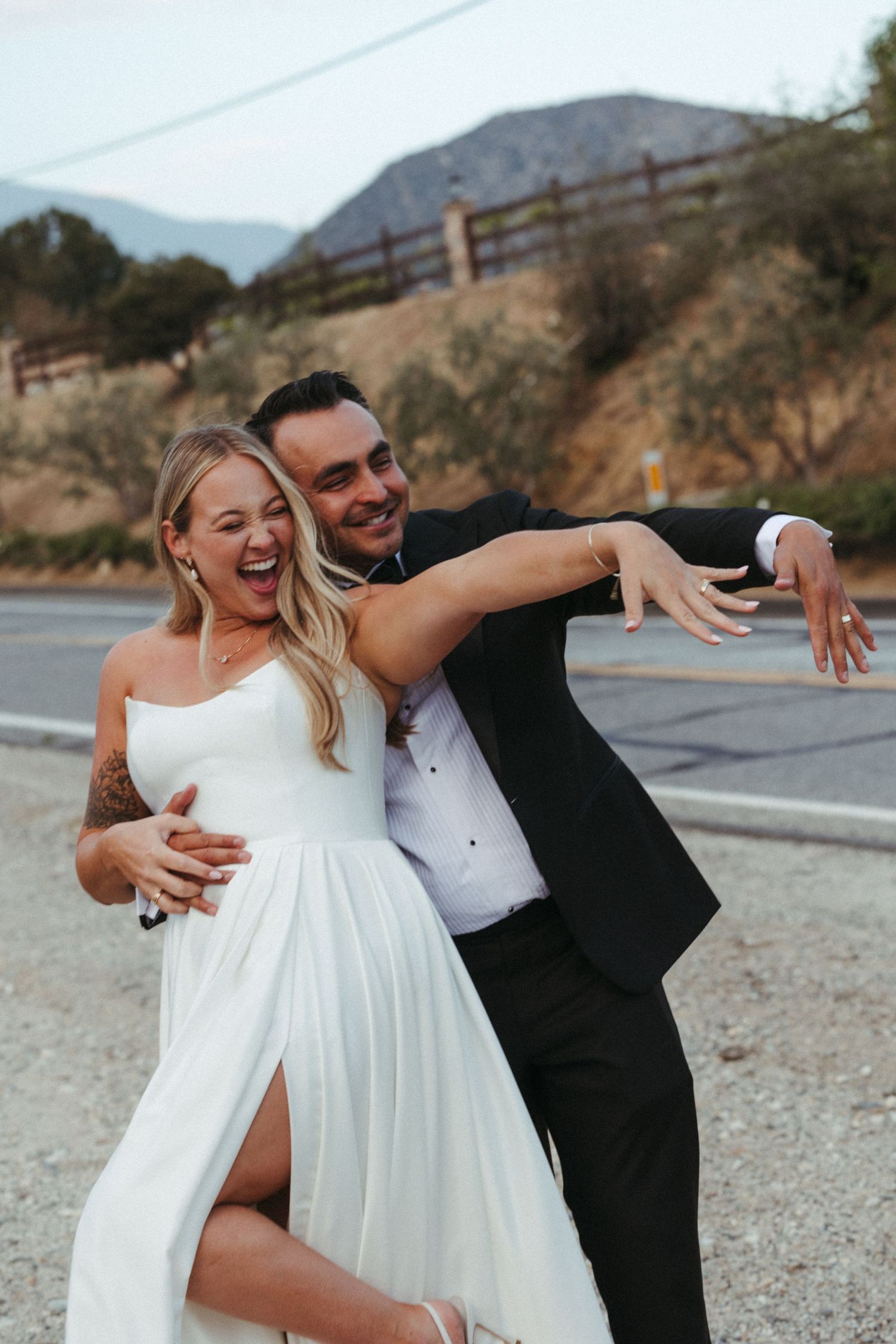Joyful bride and groom laughing and dancing together on roadside with mountain backdrop.
