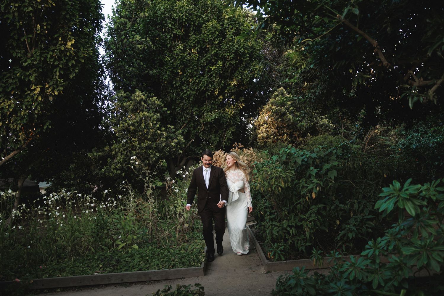 Bride and groom walk hand in hand through a lush green garden on their wedding day.