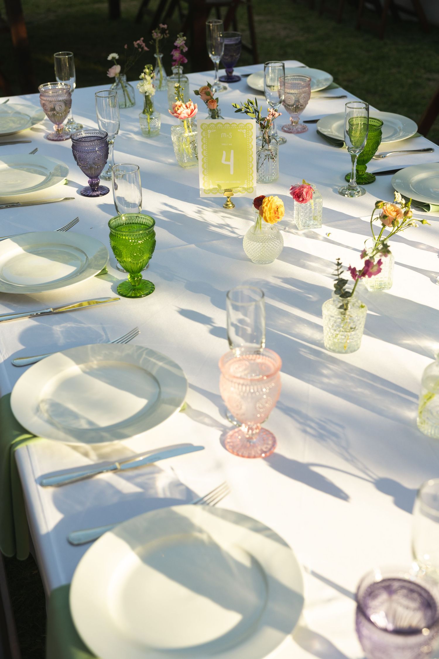 Elegant outdoor wedding table setting with colorful goblets, white plates, and small floral bud vases on a white tablecloth.