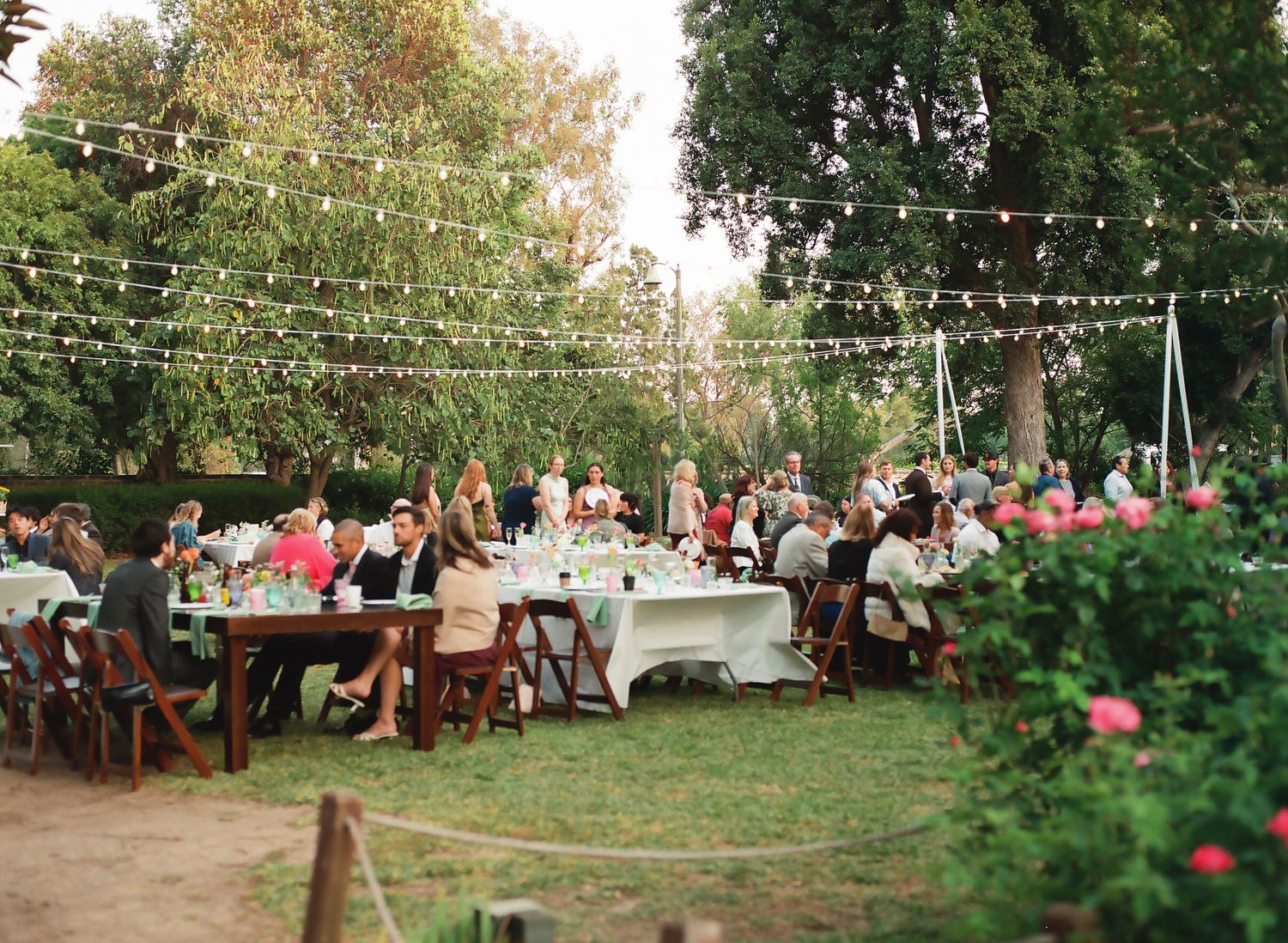 Outdoor wedding reception with string lights, long tables, and guests dining among lush greenery and pink floral arrangements.