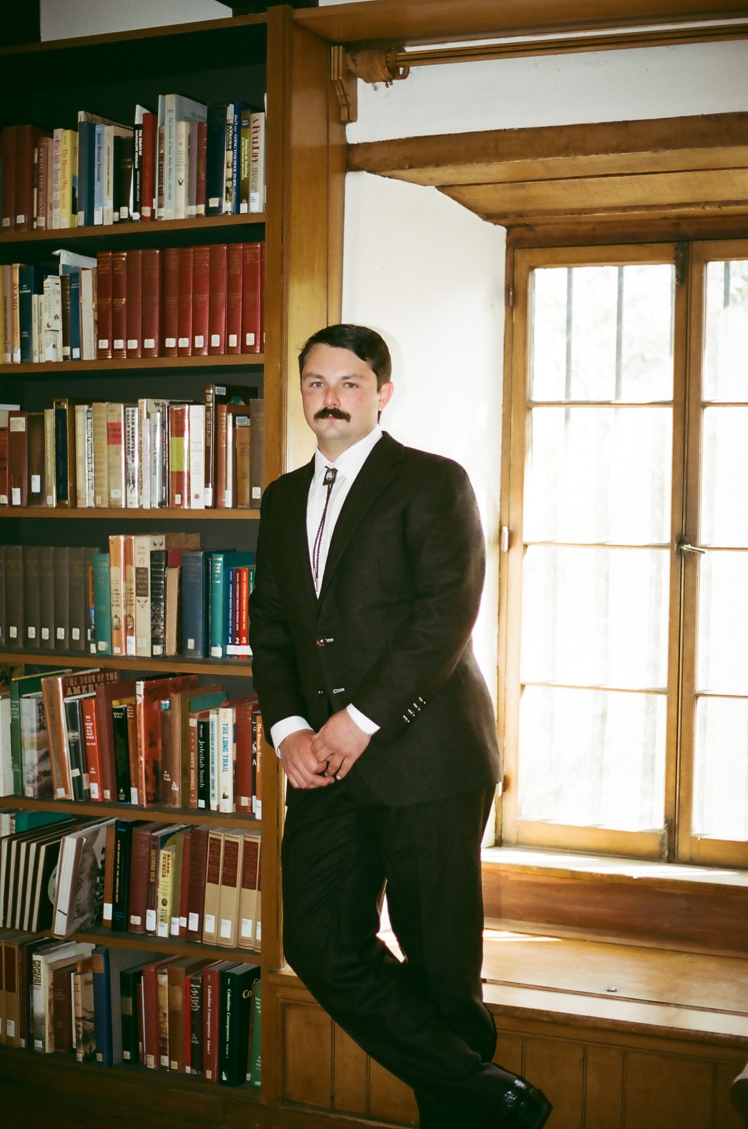 Man in dark suit with mustache leaning against bookshelf by window in classic library setting.