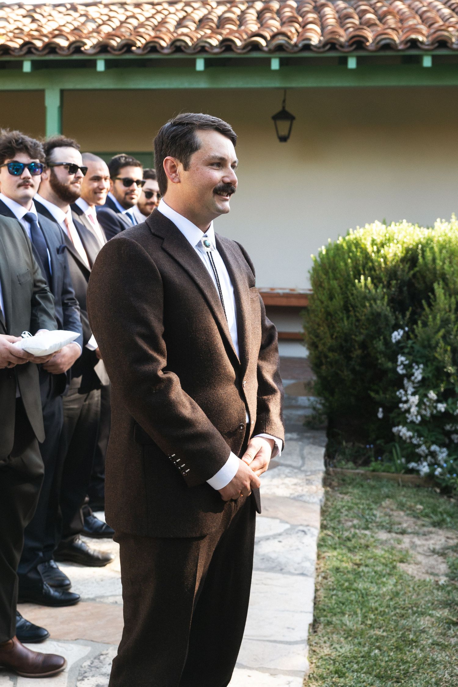 Man in brown suit with mustache smiling outdoors at formal event with group of men in suits behind him.
