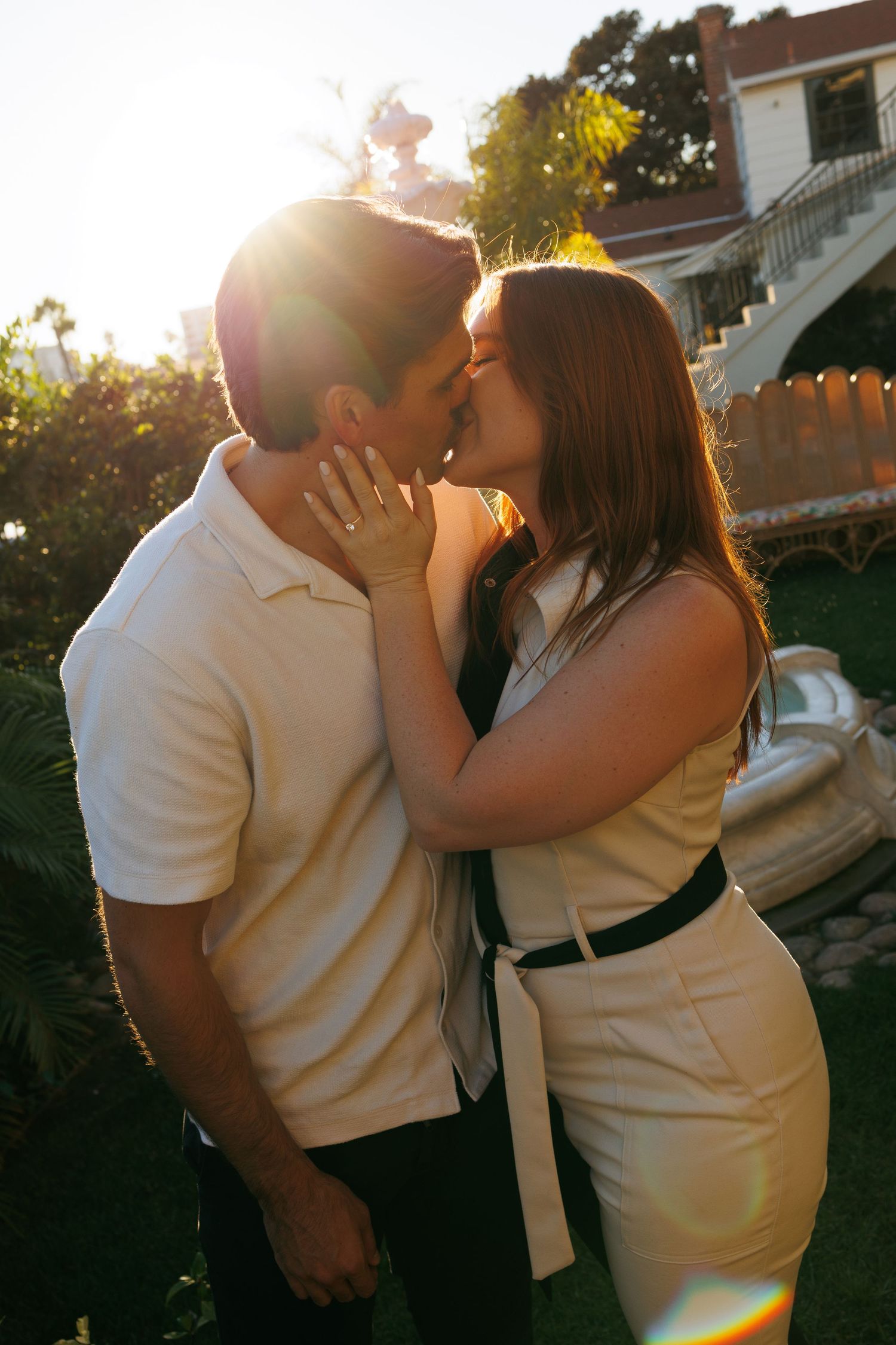 Couple sharing a kiss in golden sunlight in a lush garden, woman in white dress with black belt.