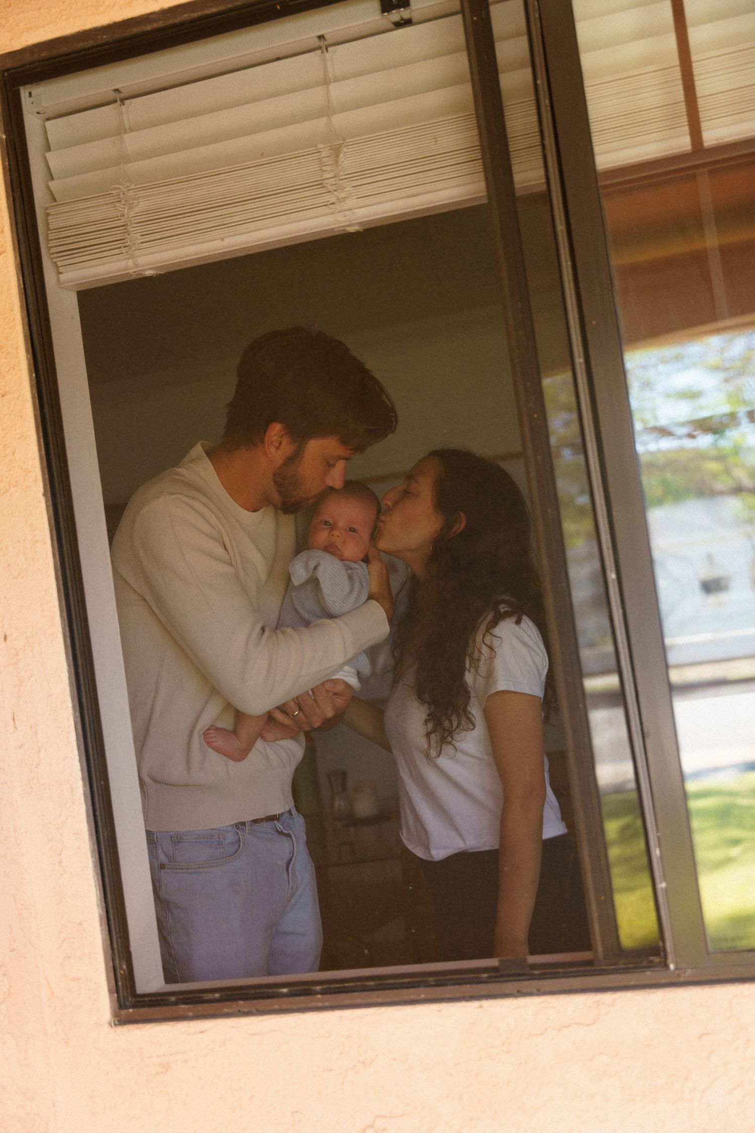 A couple shares a tender moment with their newborn baby, viewed through a window screen.