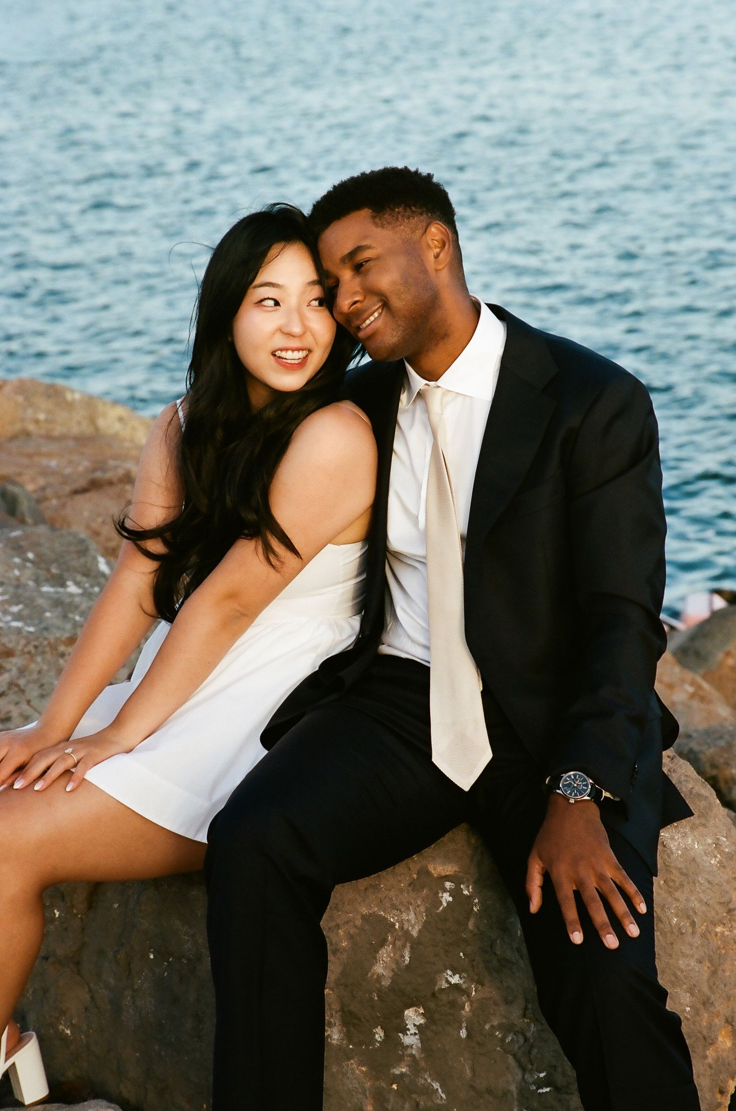 Couple in formal wear sits on rocks by water, sharing a joyful intimate moment.