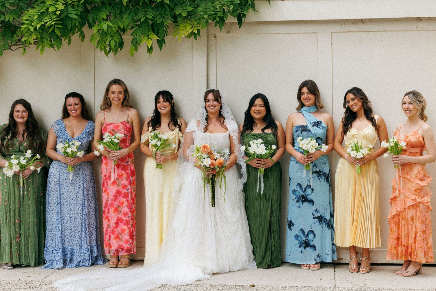 Nine bridesmaids in colorful mismatched floral dresses holding bouquets stand with bride in white gown against cream wall.