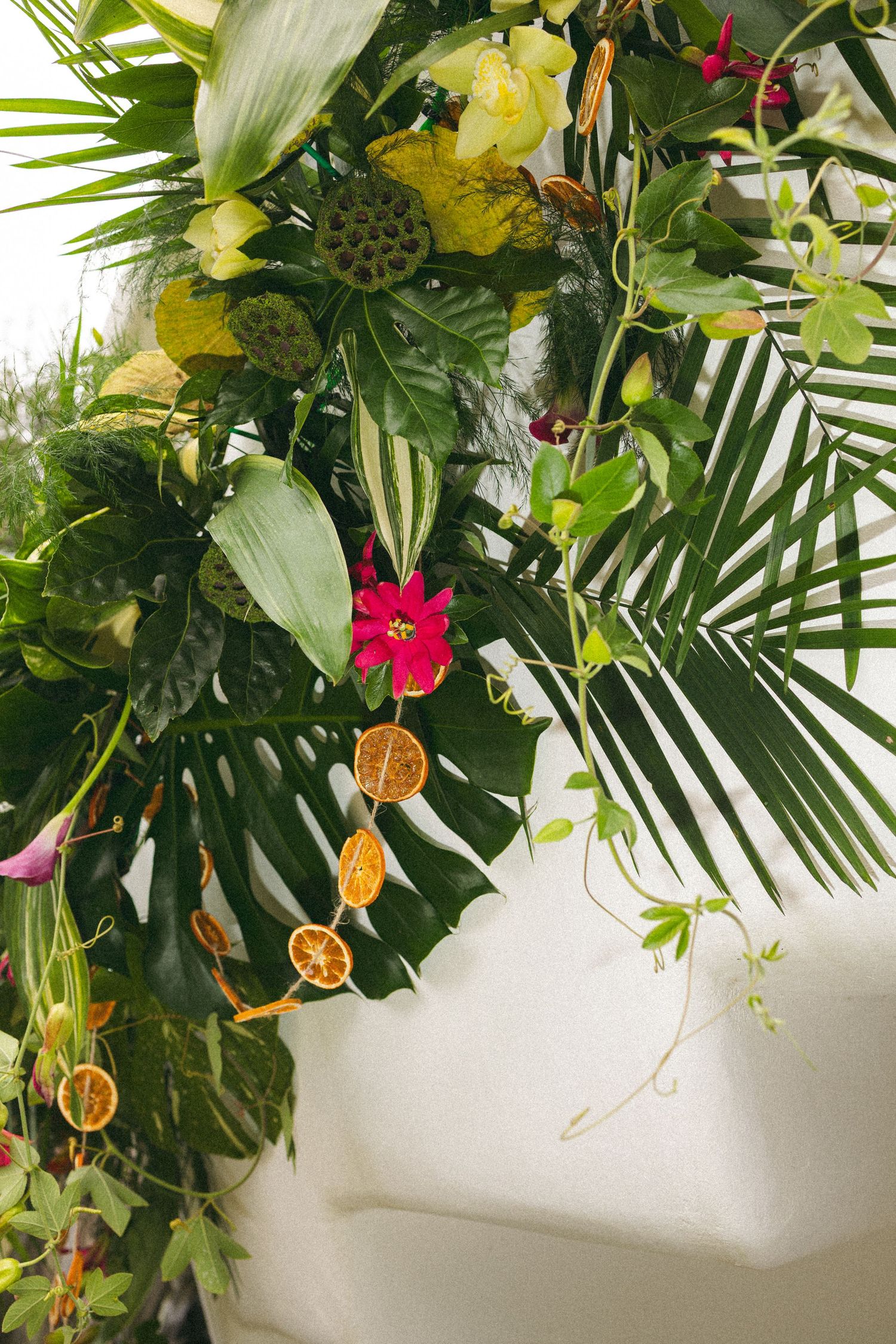 Tropical floral arrangement with monstera leaves, dried orange slices, and vibrant flowers on a white wall.