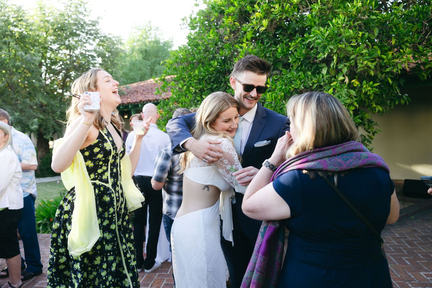 Guests laughing and hugging at an outdoor wedding reception, with a man in sunglasses embracing a woman in white.